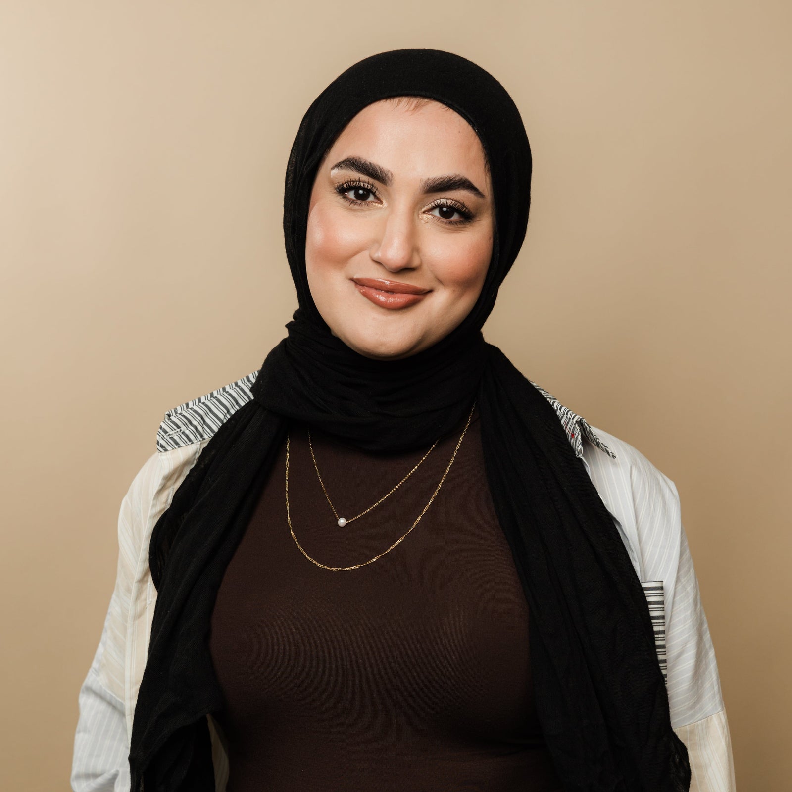 Woman wearing layered gold necklaces with a twisted chain and pearl pendant, styled with a brown top, black hijab, and striped detail shirt.