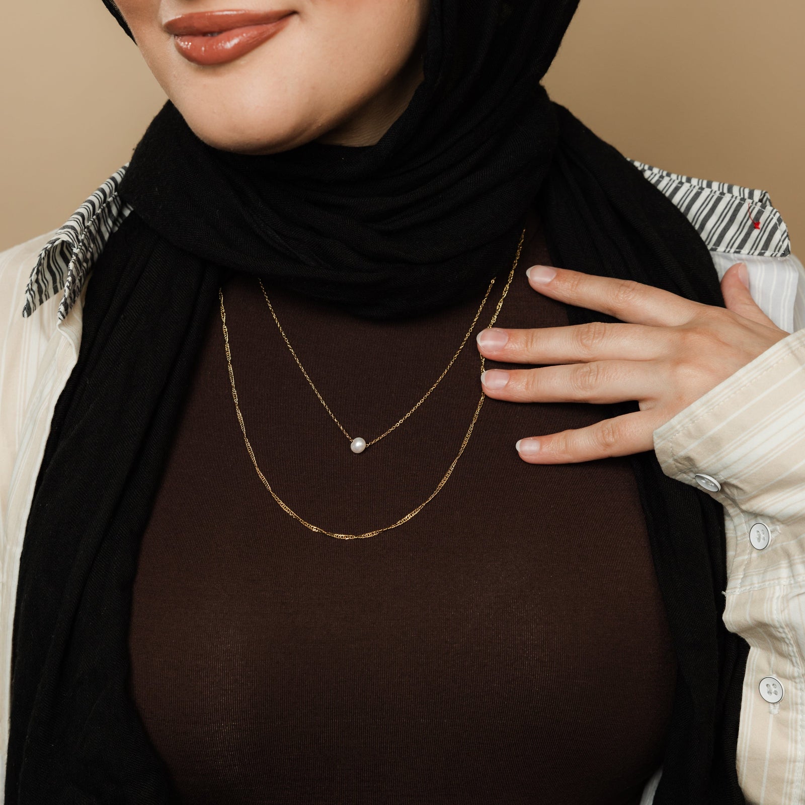 Woman wearing layered gold necklaces with a rope chain and a pearl pendant chain, styled with a brown top and black hijab, hand resting near the collar.