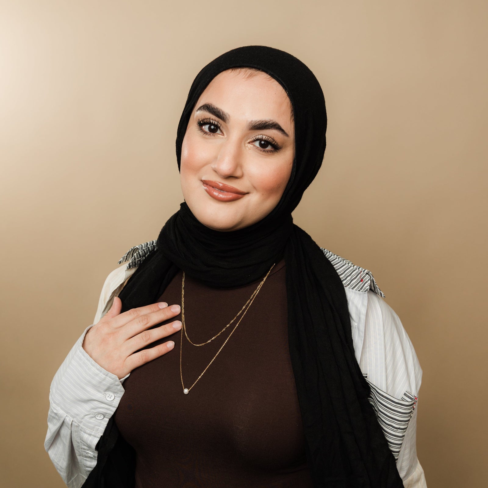 Woman wearing layered gold necklaces with a twisted chain and pearl pendant, styled with a brown top, black hijab, and striped detail shirt, hand resting near the necklace.