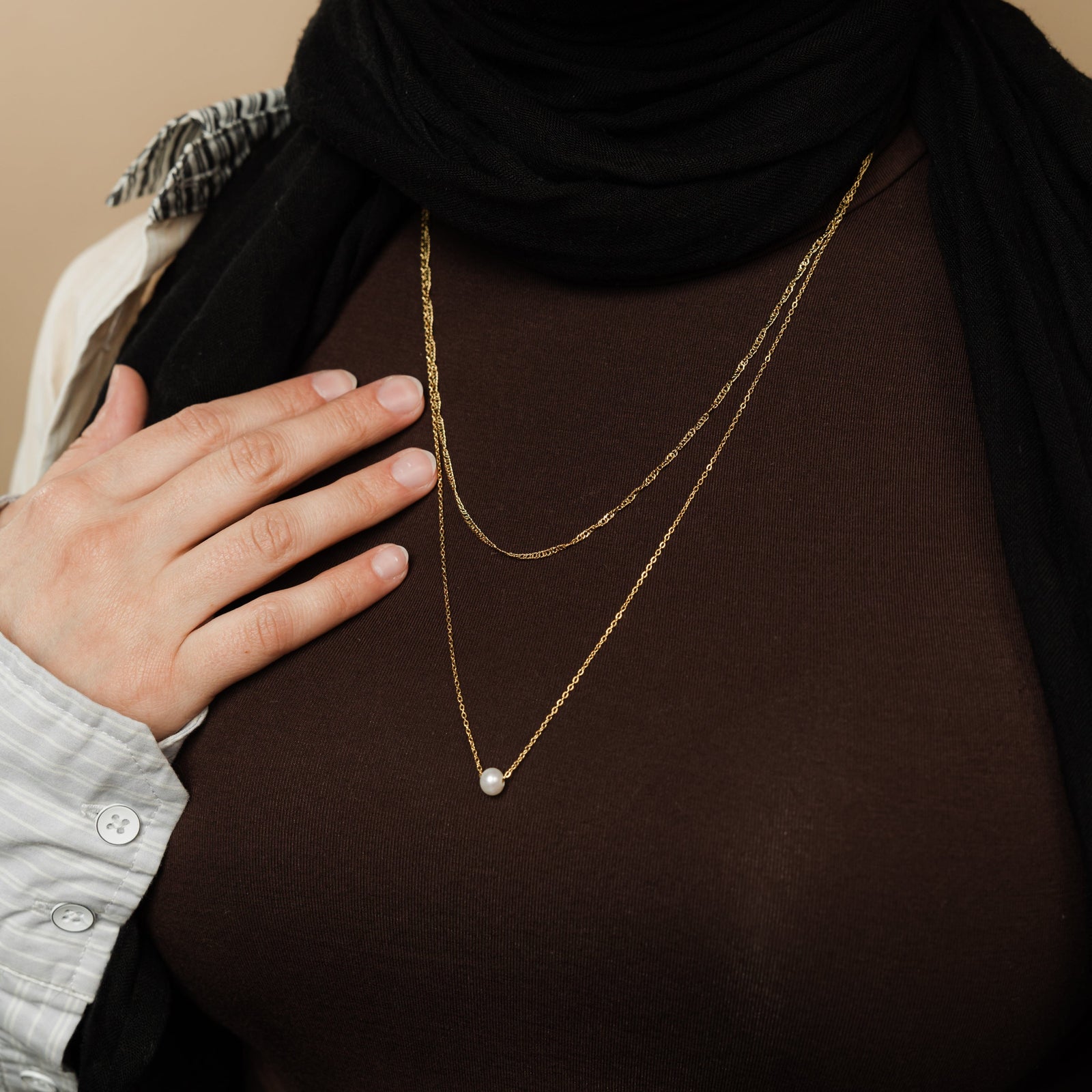 Close-up of layered gold necklaces with a twisted chain and a pearl pendant chain, worn over a brown top with hand resting near the chest.
