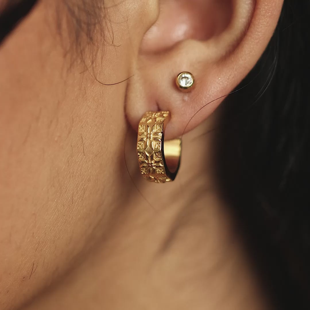 Close up photo of the model's left ear wearing the gold earrings and a simple circle stud earrings. Closer look at the tatreez design of the gold Tatreez Hoop Earrings. Model moves their head to show both pair of the gold hoop earrings.