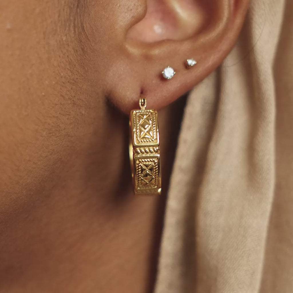 Close up of the gold Damascus Tile Earrings as worn by the model on their left ear. The model touches the gold earring with their index finger. A closer look to the designs of the gold hook earrings.