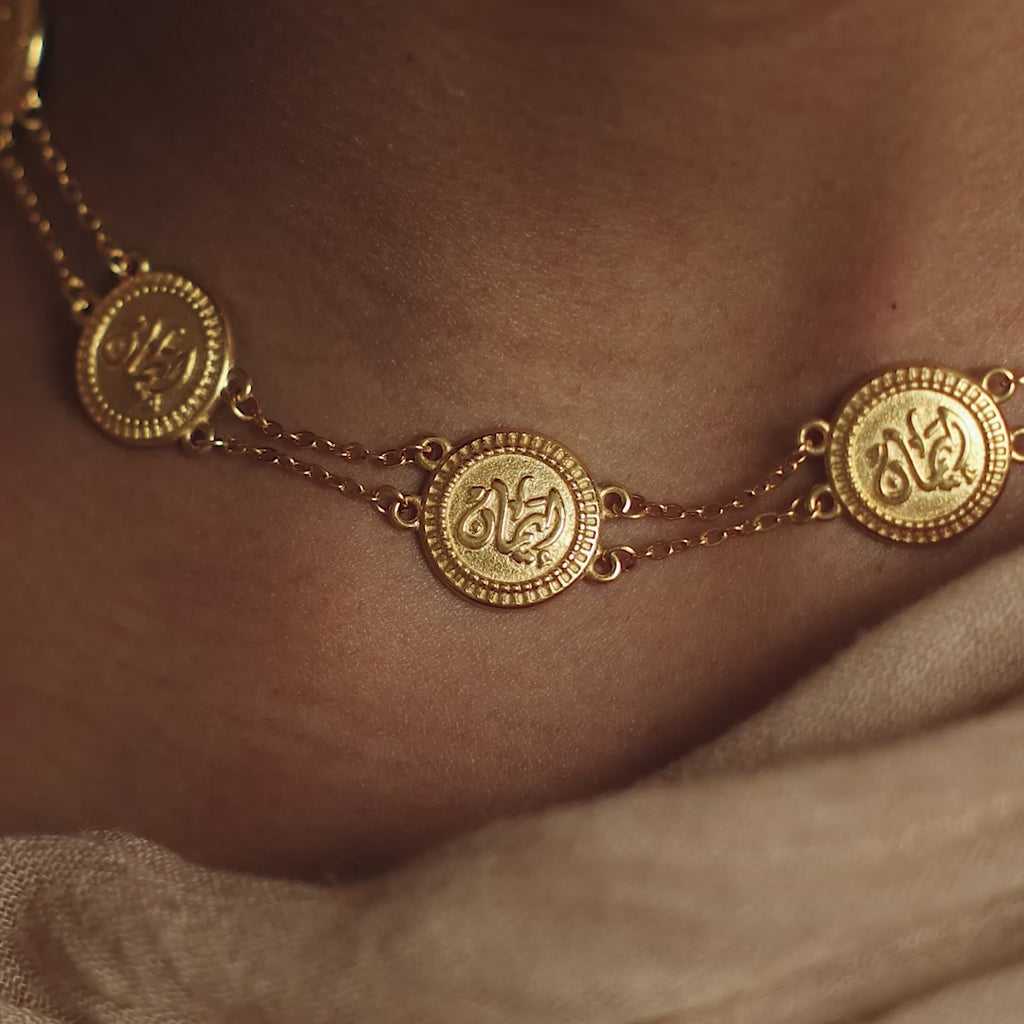 Close up of the gold Iman | Faith Coin Necklace, showing it's inscription. Model moves around to show the gold necklace on their neck. Model smiles at the camera, wearing the gold Iman | Faith Coin Necklace.