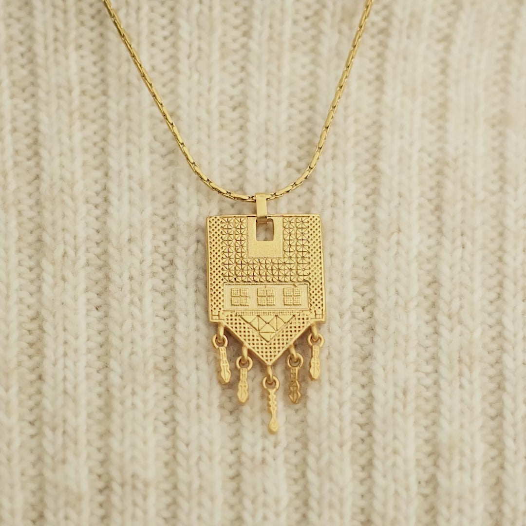 Close up of the gold Raining Tatreez Necklace against a light fabric. Model adjusts the gold necklace. Camera moves, focused on the pendant of the the necklace.