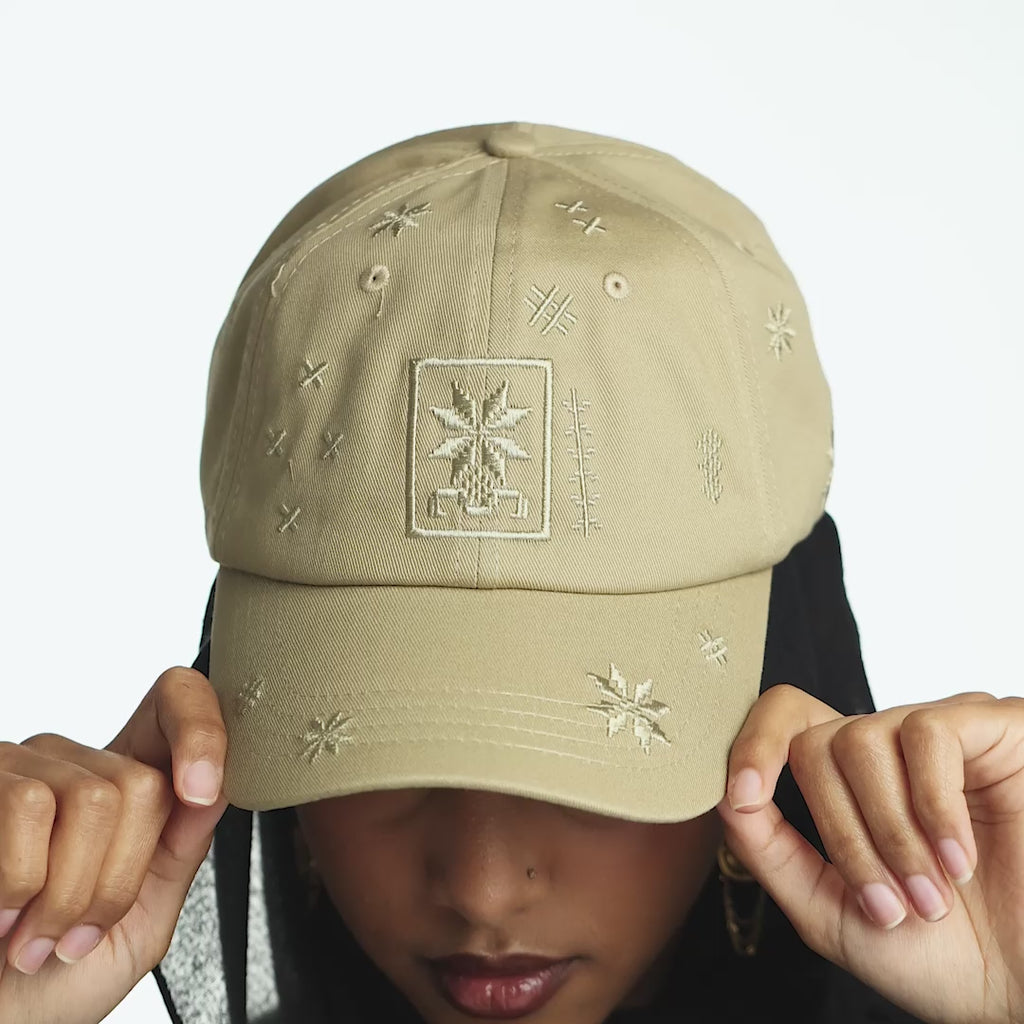 Model holds on to the cap, looking at the camera. Close up of the embroidery design of the beige cap. Model demonstrates how to adjust the clasp of the cap. Model wears the cap on their head.
