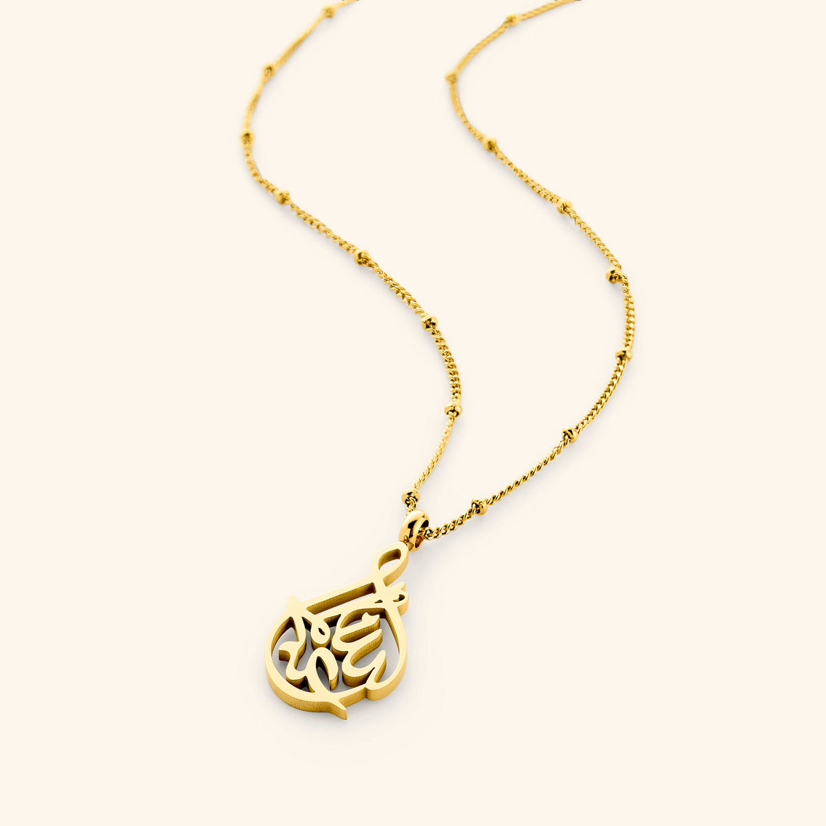 Flat lay of an 18k gold plated necklace on a light background. The necklace features calligraphy pendant on a thin dotted chain.