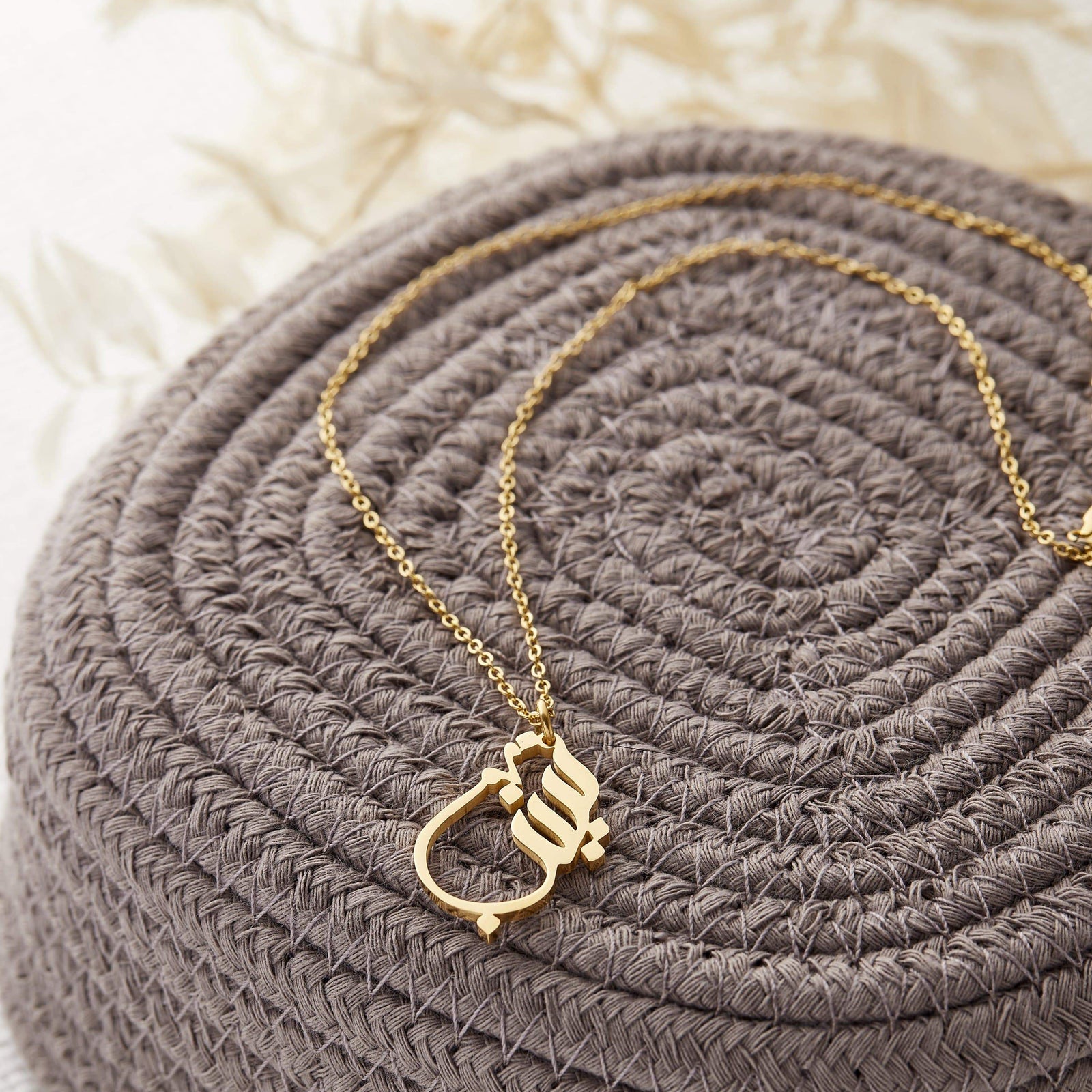 Photo of a gold Ready Calligraphy Name Necklace laid flat on a knitted fabric.