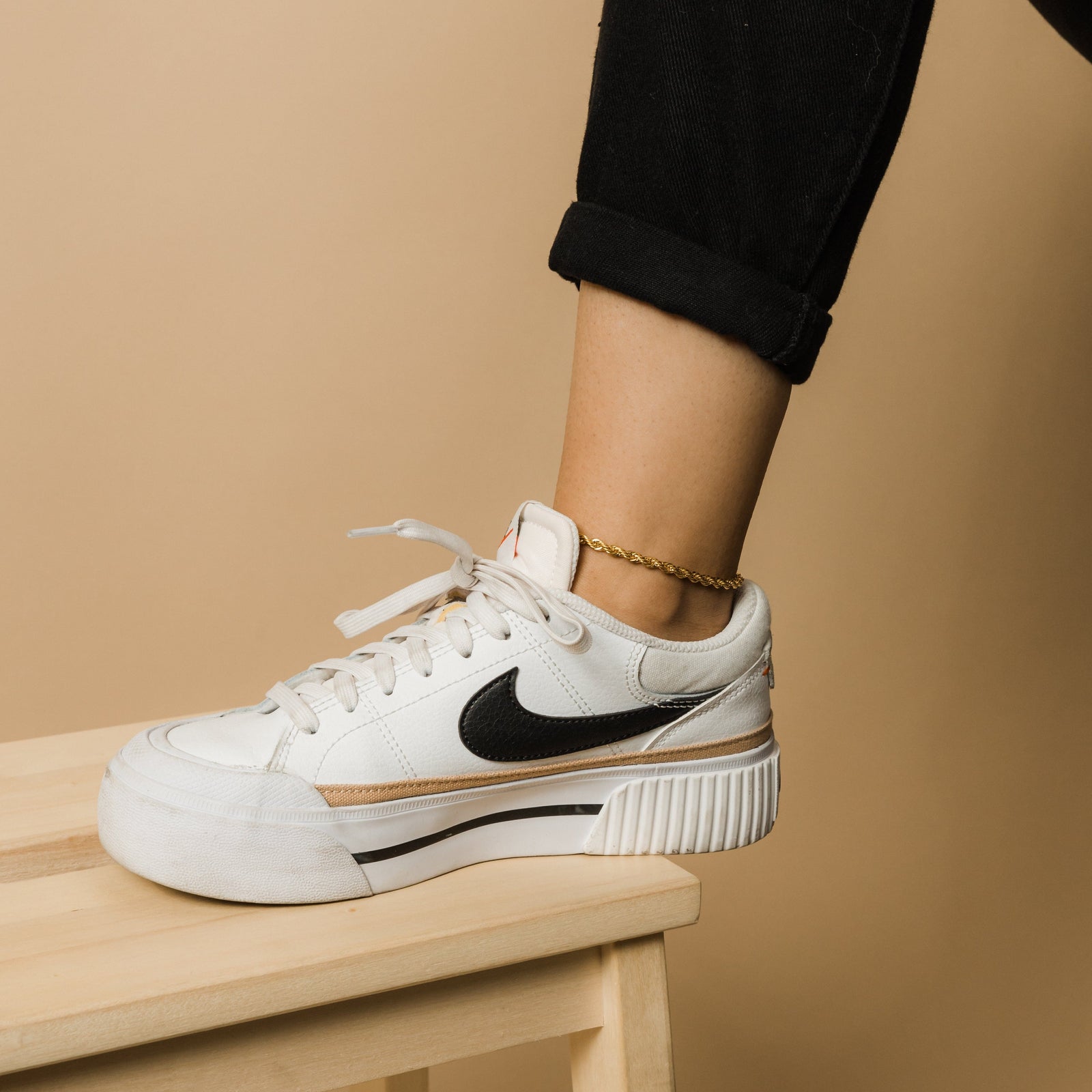 Gold rope chain anklet worn on ankle above a white sneaker, leg resting on a wooden stool.