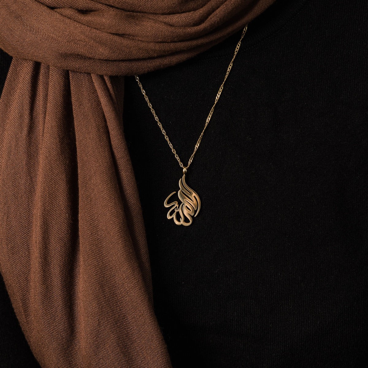 Close up of the gold Alhamdulillah Calligraphy Necklace resting against the black fabric of the model's clothing.