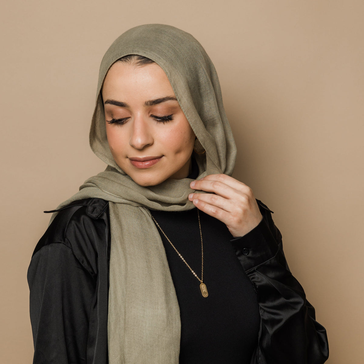 Photo of the model looking down and touching their hijab with their left hand. The model is wearing the gold Alhamdulillah Moon Necklace | Women.