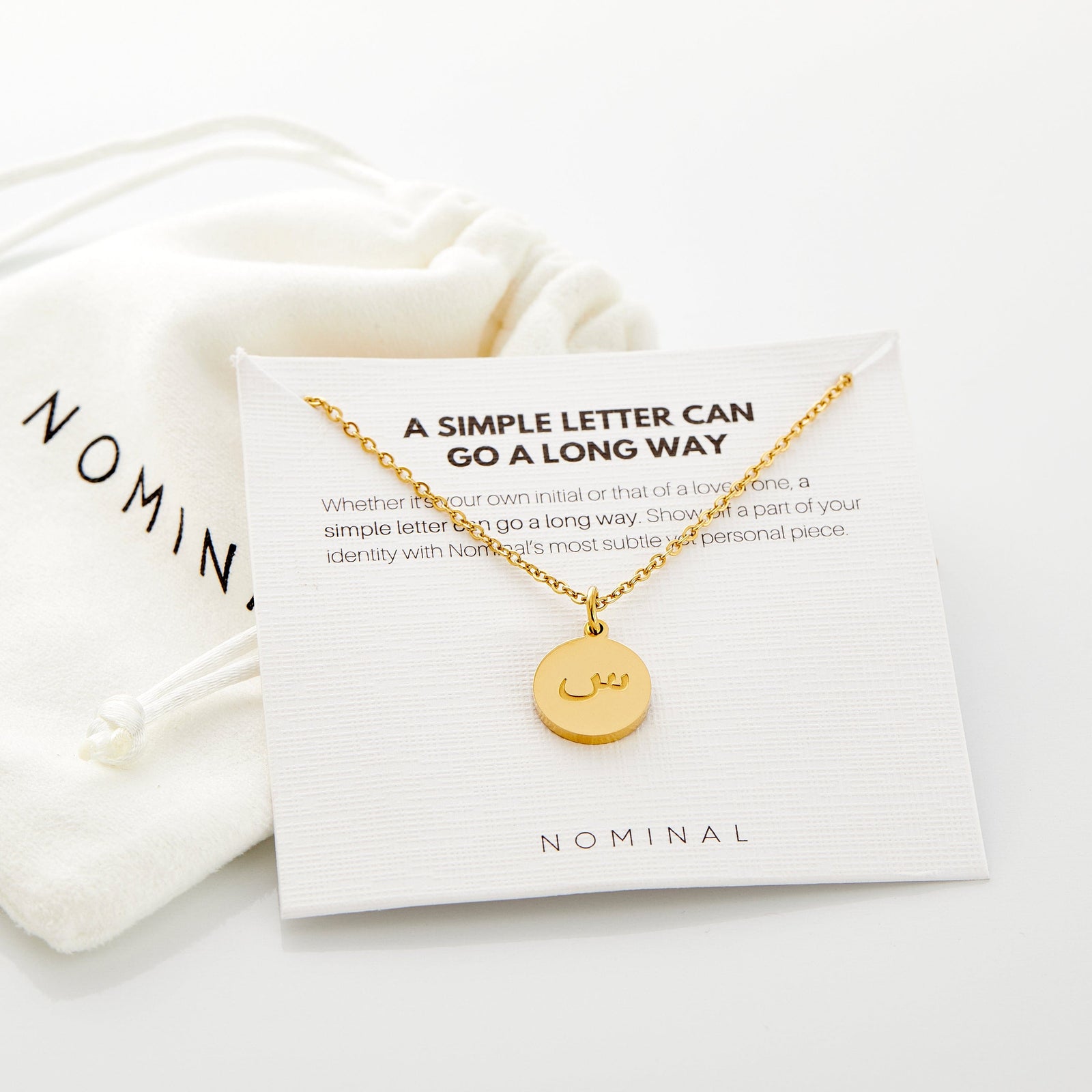 A delicate gold-plated necklace with a circular pendant. An Arabic letter is elegantly engraved in the center of the pendant. The necklace has a thin chain and is displayed on a light beige background. The specific Arabic letter is clearly visible within the circular frame of the pendant.