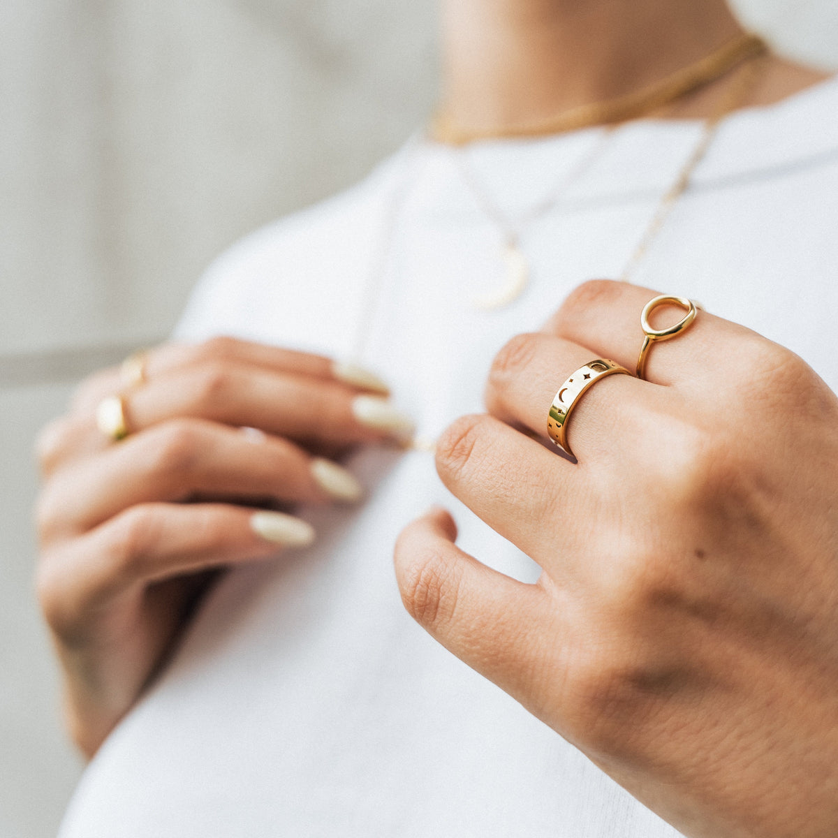 Woman’s hand wearing the gold Stargazer’s Ring with crescent moon and star cutouts, paired with an open-circle ring, styled with layered gold necklaces over a white top.