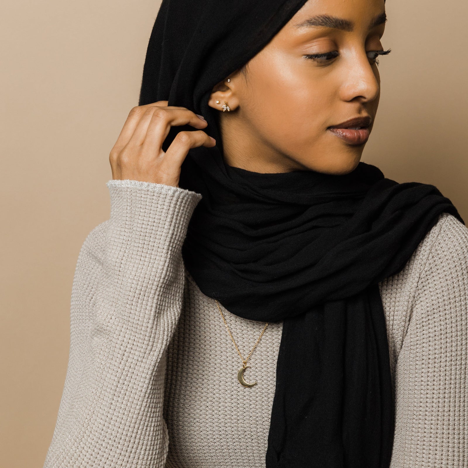 Photo of the model looking to her left side and touching their hijab. The model is wearing the gold Crescent Moon Necklace.