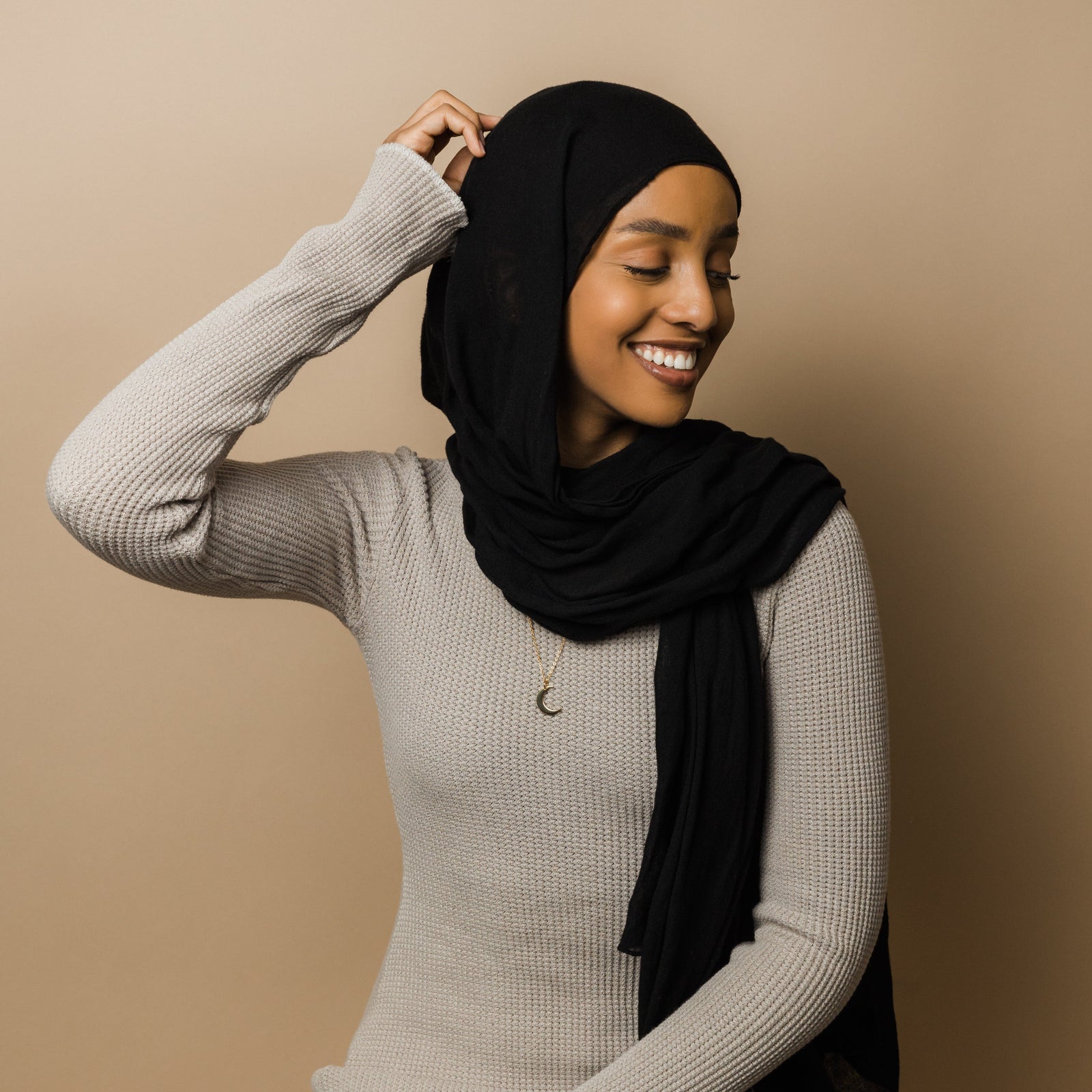 Photo of the model touching the top of their head over their black hijab. The model is wearing the gold Crescent Moon Necklace.