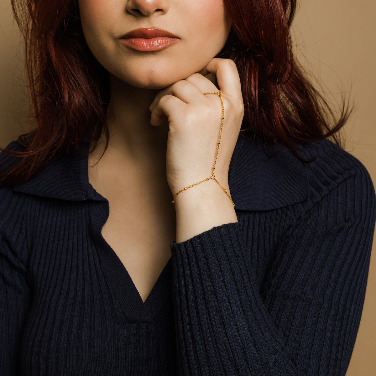 Woman wearing a gold dotted hand chain, styled with a navy ribbed long-sleeve top, resting her hand near her chin.