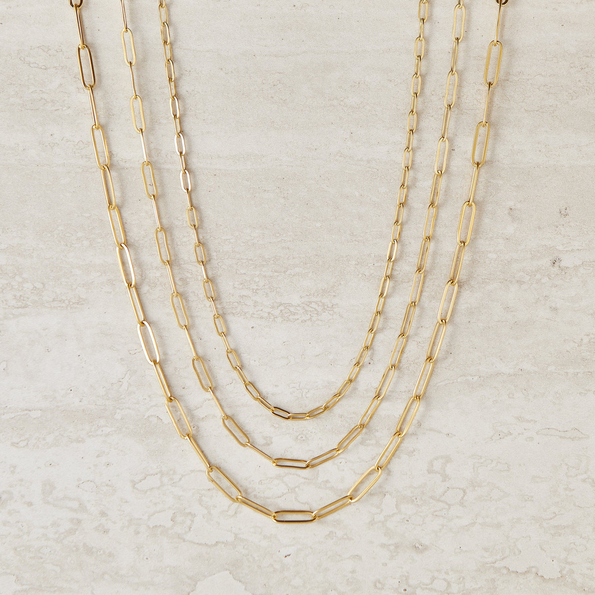 Three gold link chain necklaces of varying thicknesses and lengths displayed against a light beige background.