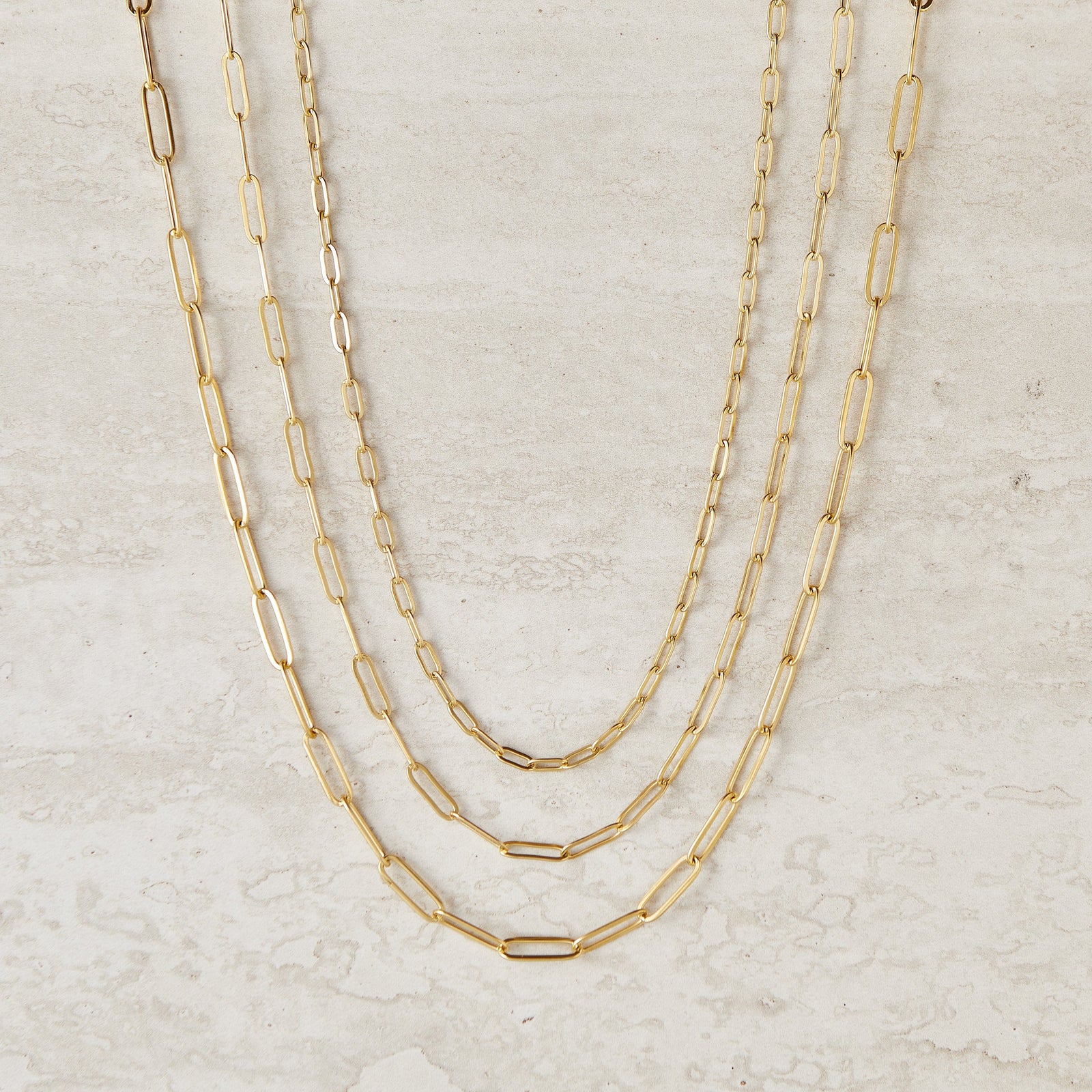 Three gold link chain necklaces of varying thicknesses and lengths displayed against a light beige background.