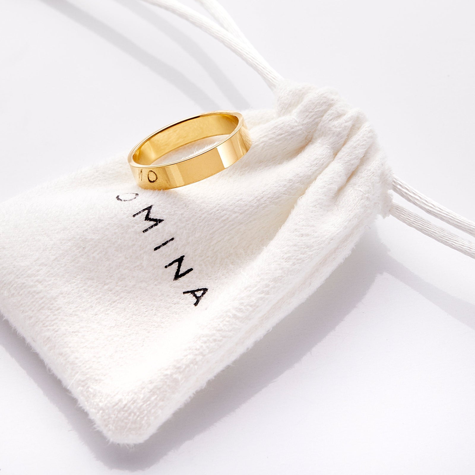 Gold flat band ring resting atop a white drawstring pouch with 'Nominal' branding.