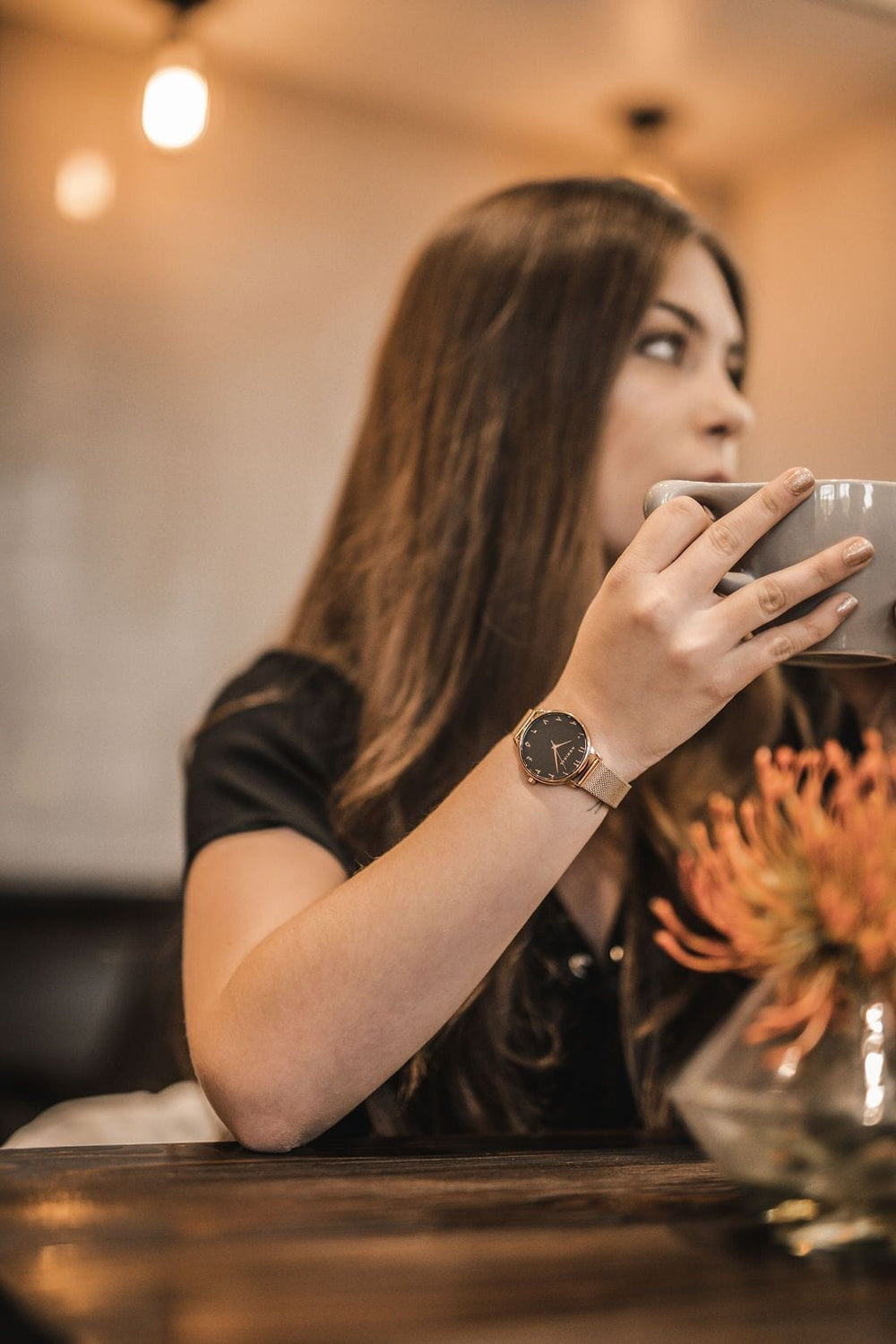 Close-up of a gold mesh strap watch with minimalist Arabic numerals worn on the wrist of a woman who is sipping tea from a grey cup, she is wearing a dark shirt, and it's positioned against a neutral background. 