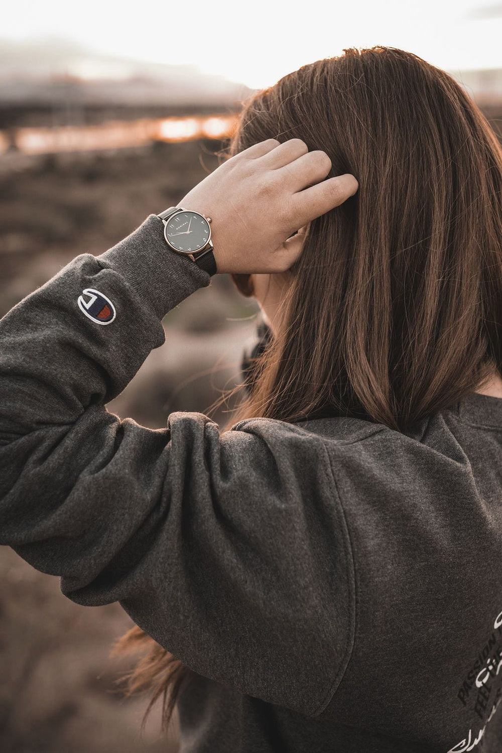 Close-up of a black strap watch with gold details and with Arabic numerals, worn on the wrist of a woman with a grey sweater holding her hand against her head in a natural blurred background. 