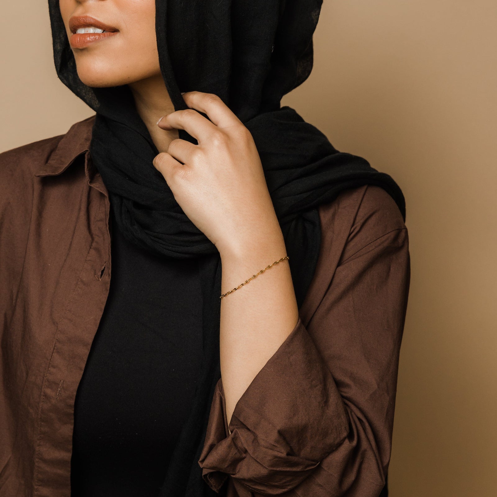 A woman in brown shirt and black hijab styling the gold flutter bracelet on wrist.