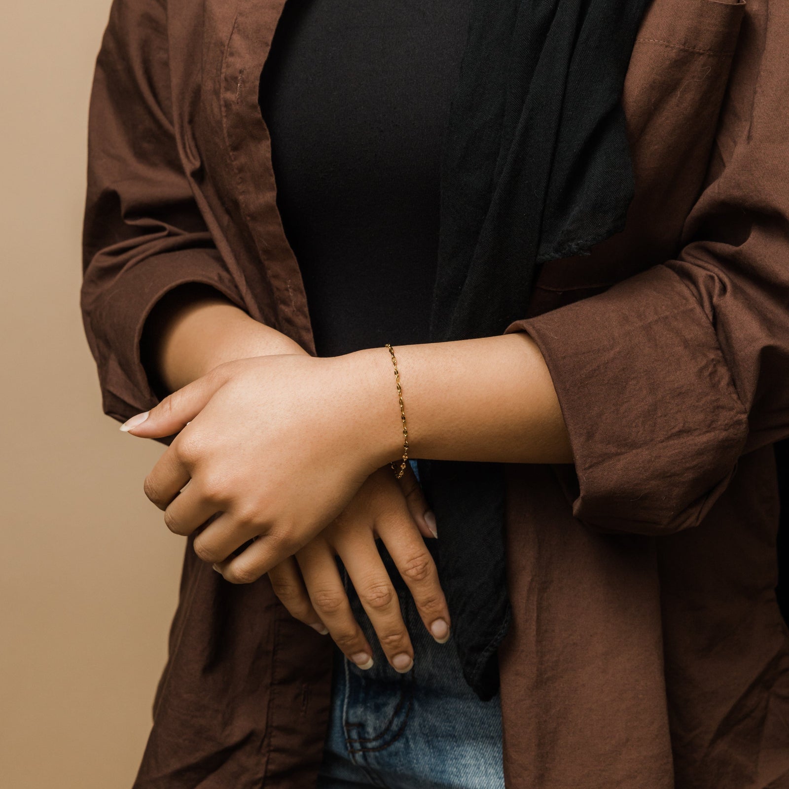 Gold flutter bracelet styled on wrist across the body, paired with black shirt, brown button-down and denim pants.