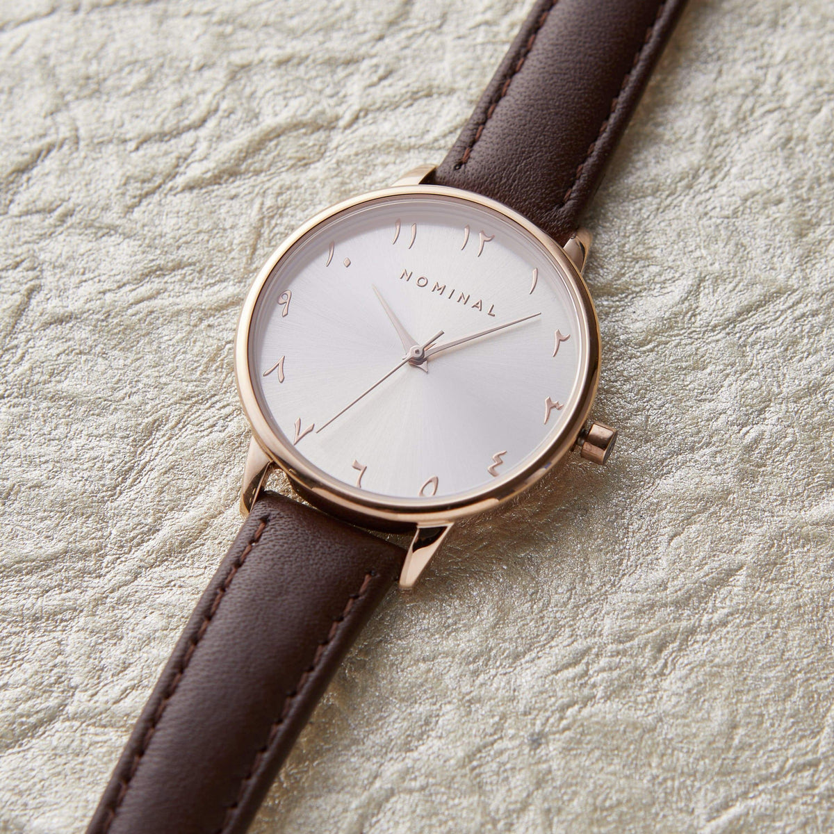 Brown leather  trap watch with with background and rose gold details, with minimalist Arabic numerals against a neutral textured background.