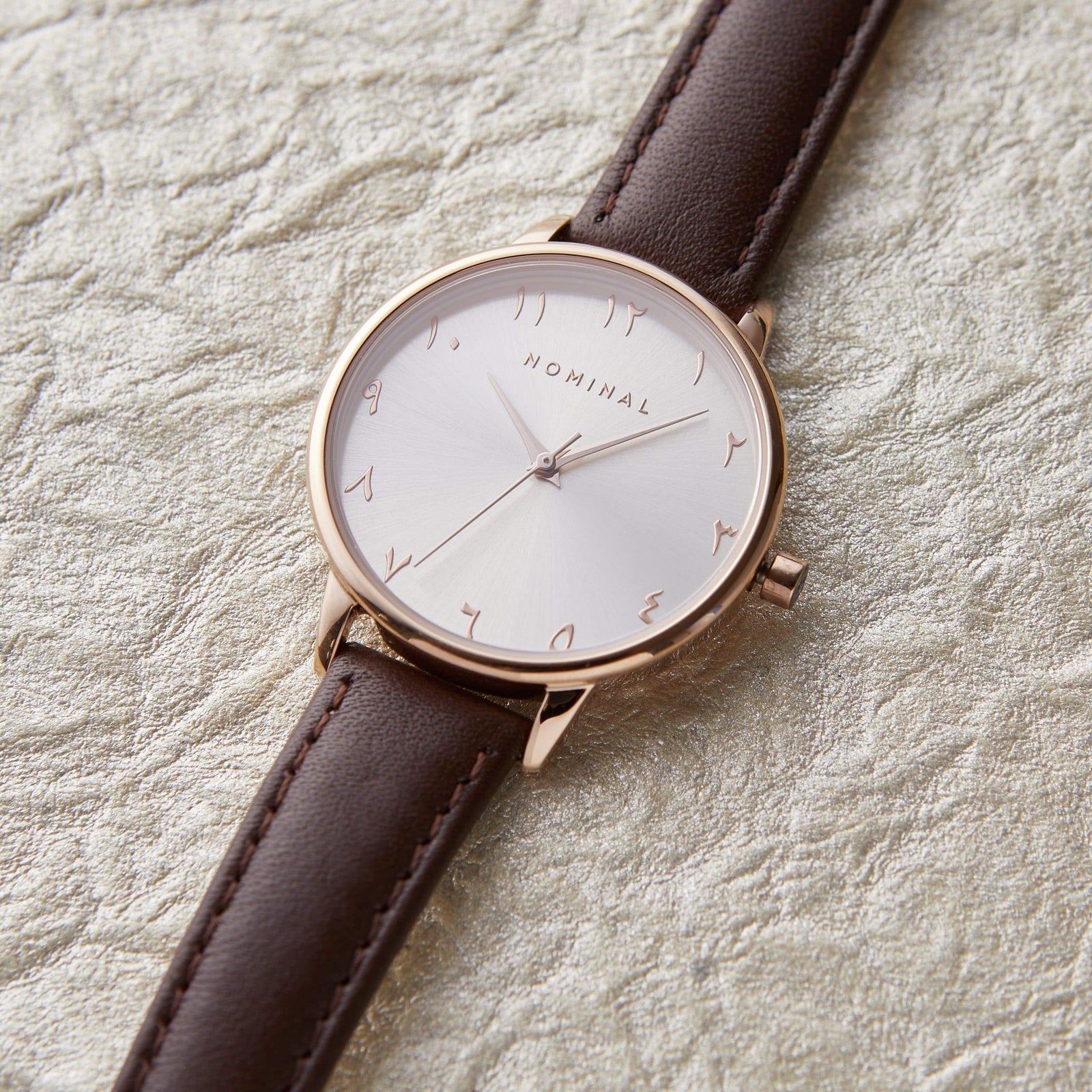 Brown leather  trap watch with with background and rose gold details, with minimalist Arabic numerals against a neutral textured background.