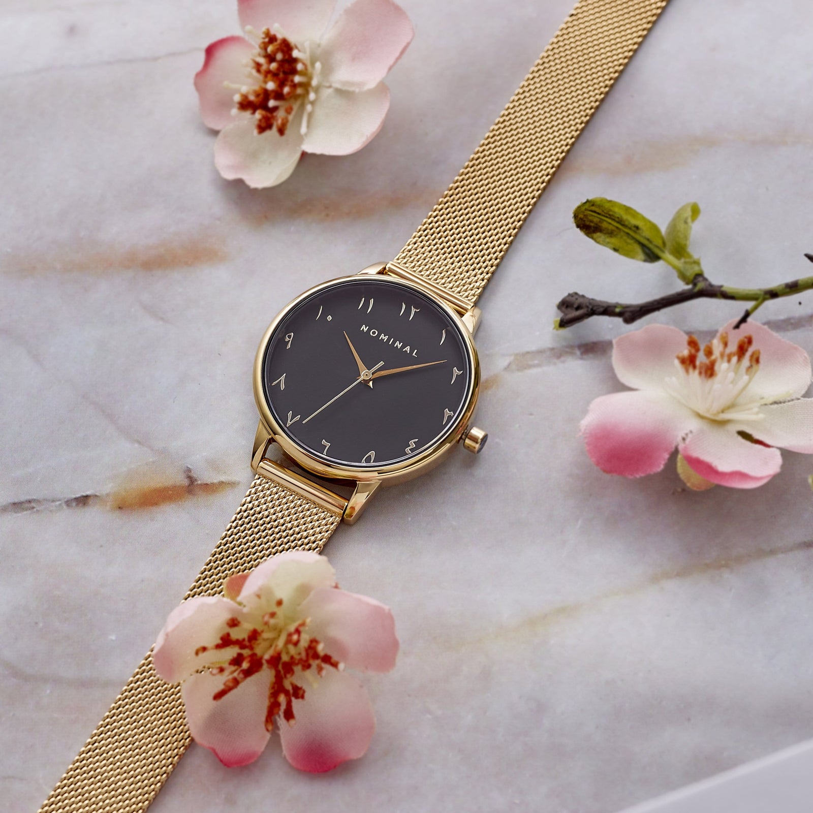 Gold mesh strap watch against a grey background with pink and white with minimalist Arabic numerals in gold. 