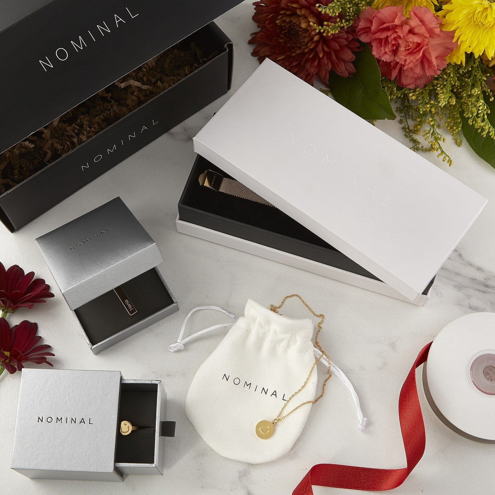 Gold jewelry pieces including a mesh strap watch, ring, and necklace with crescent charm, elegantly arranged in Nominal branded boxes and pouches on a marble background with flowers.
