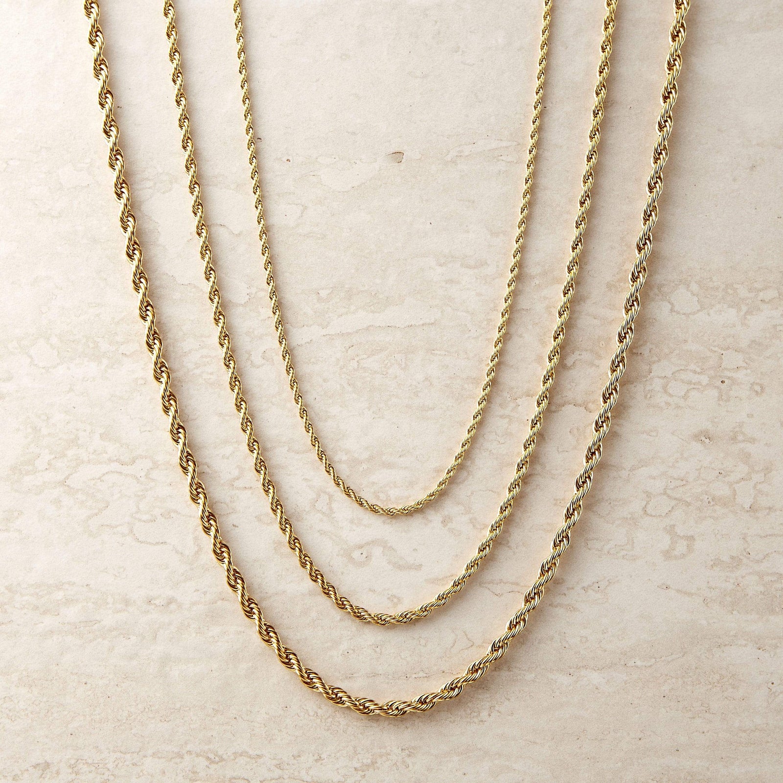 Three gold rope chain necklaces of varying thicknesses and lengths layered together on a beige background.