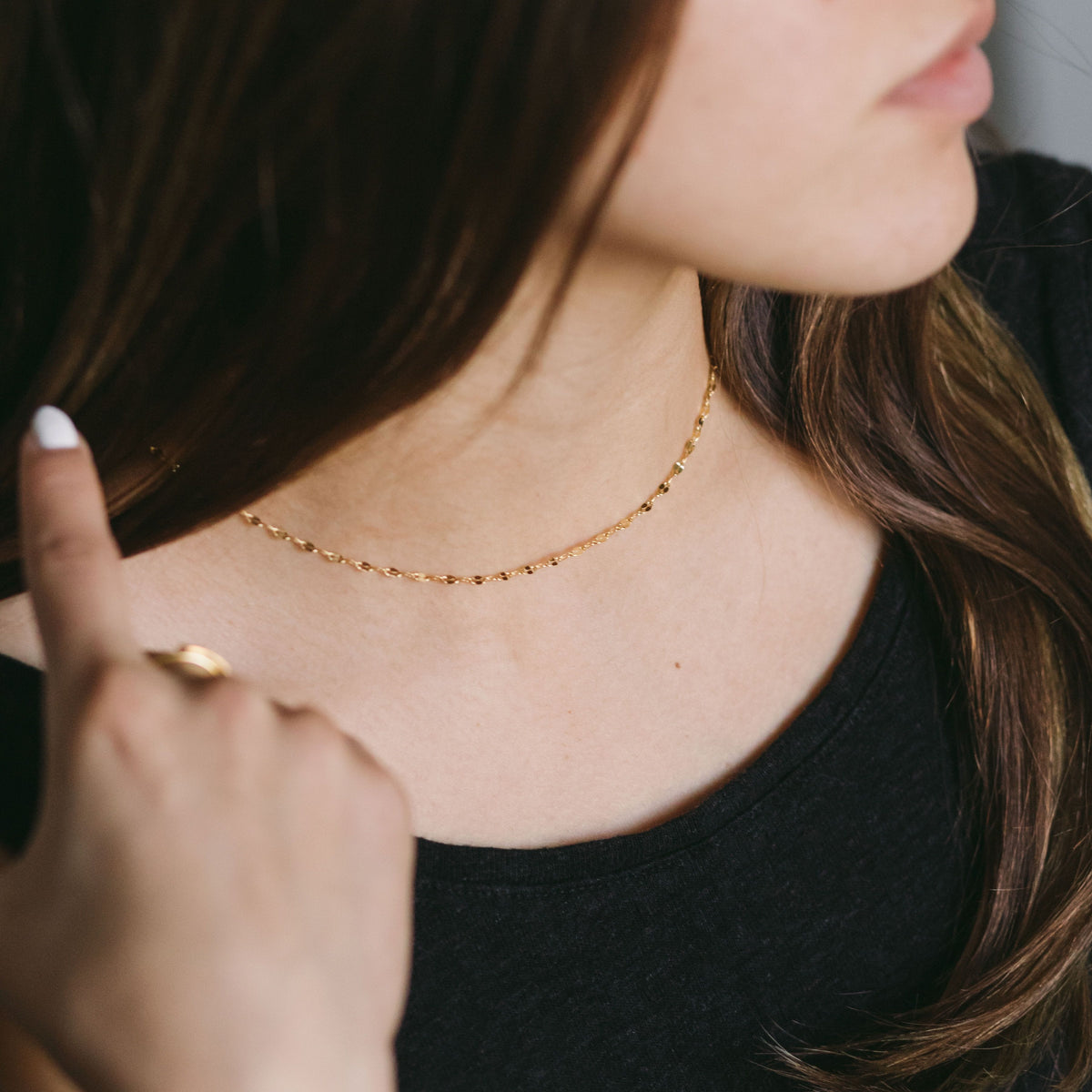 Woman wearing a gold flutter chain necklace styled with a black top, touching her hair.