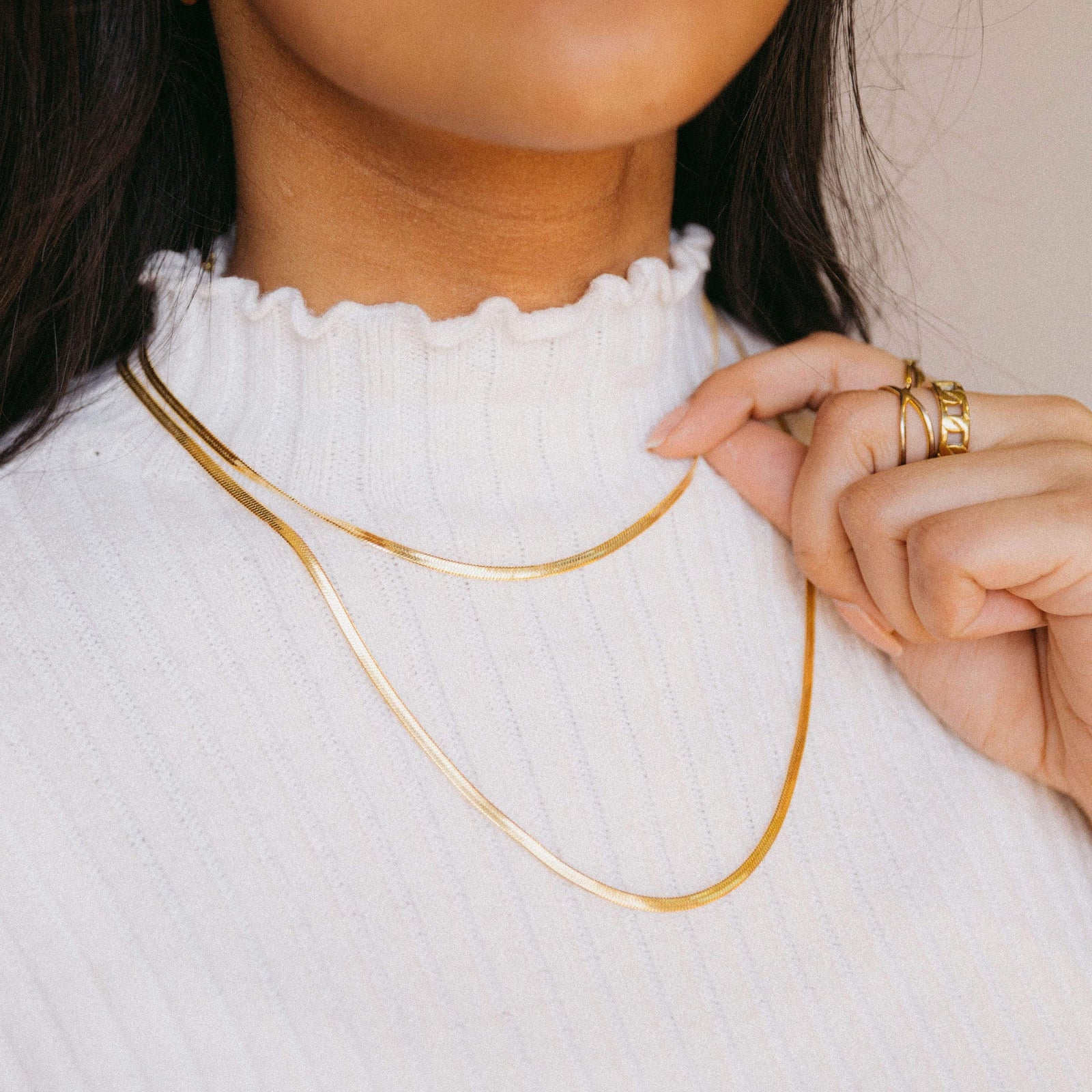 Model wearing two gold herringbone chains over a white ribbed turtleneck, holding one chain with her left hand.