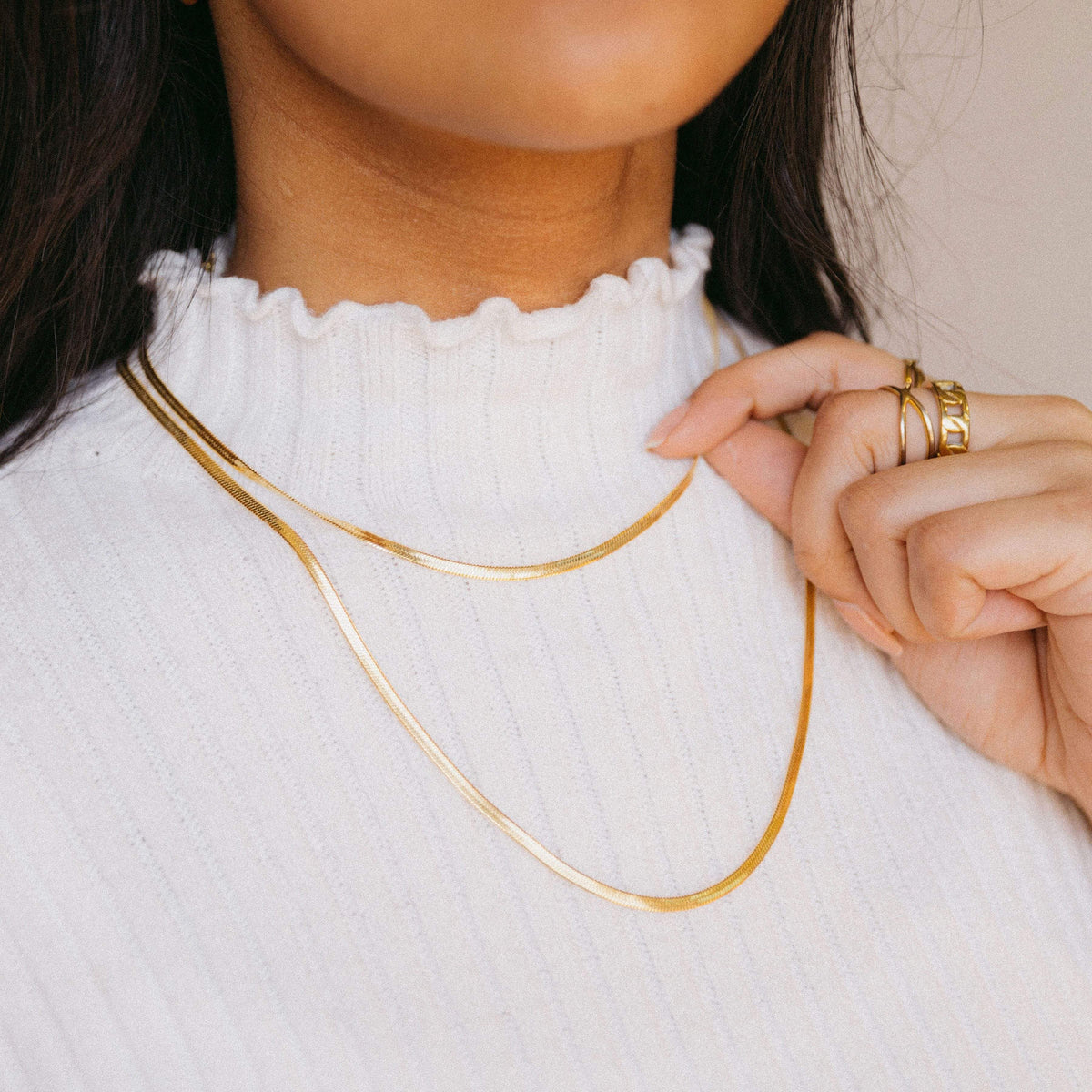 Woman wearing two layered gold herringbone necklaces over a ribbed white high-neck top, hand gently touching the shorter chain.