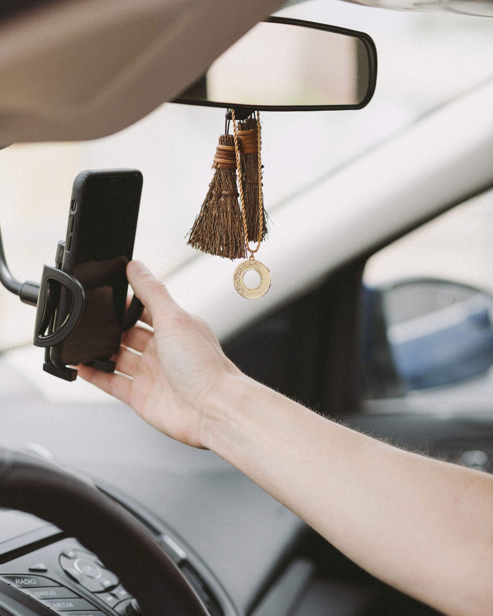 Gold Duaa Al-Safar car hanger hanging from a rearview mirror alongside tassel ornaments, hand holding a phone in the foreground.