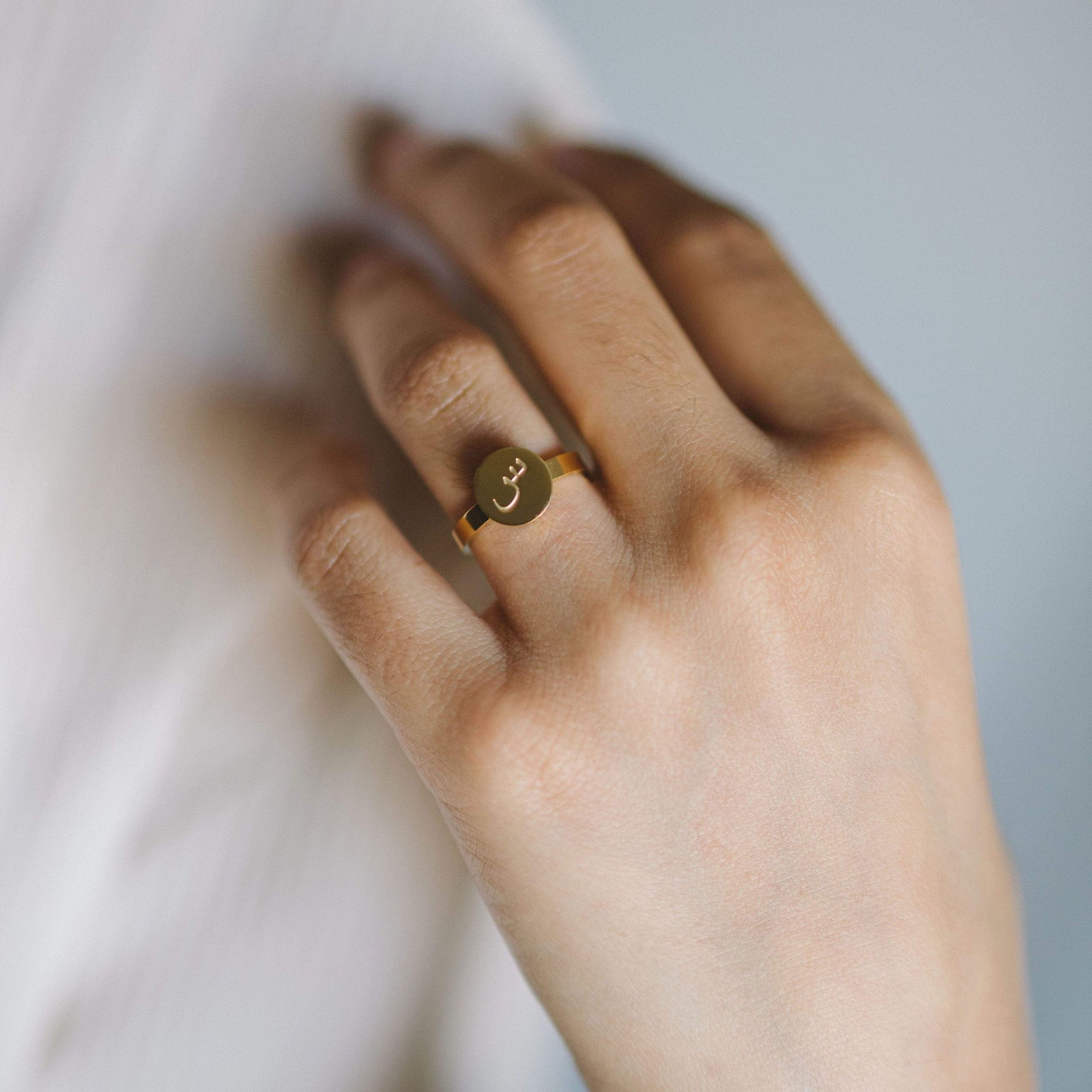 Hand wearing a gold ring with a circular face engraved with an Arabic letter.