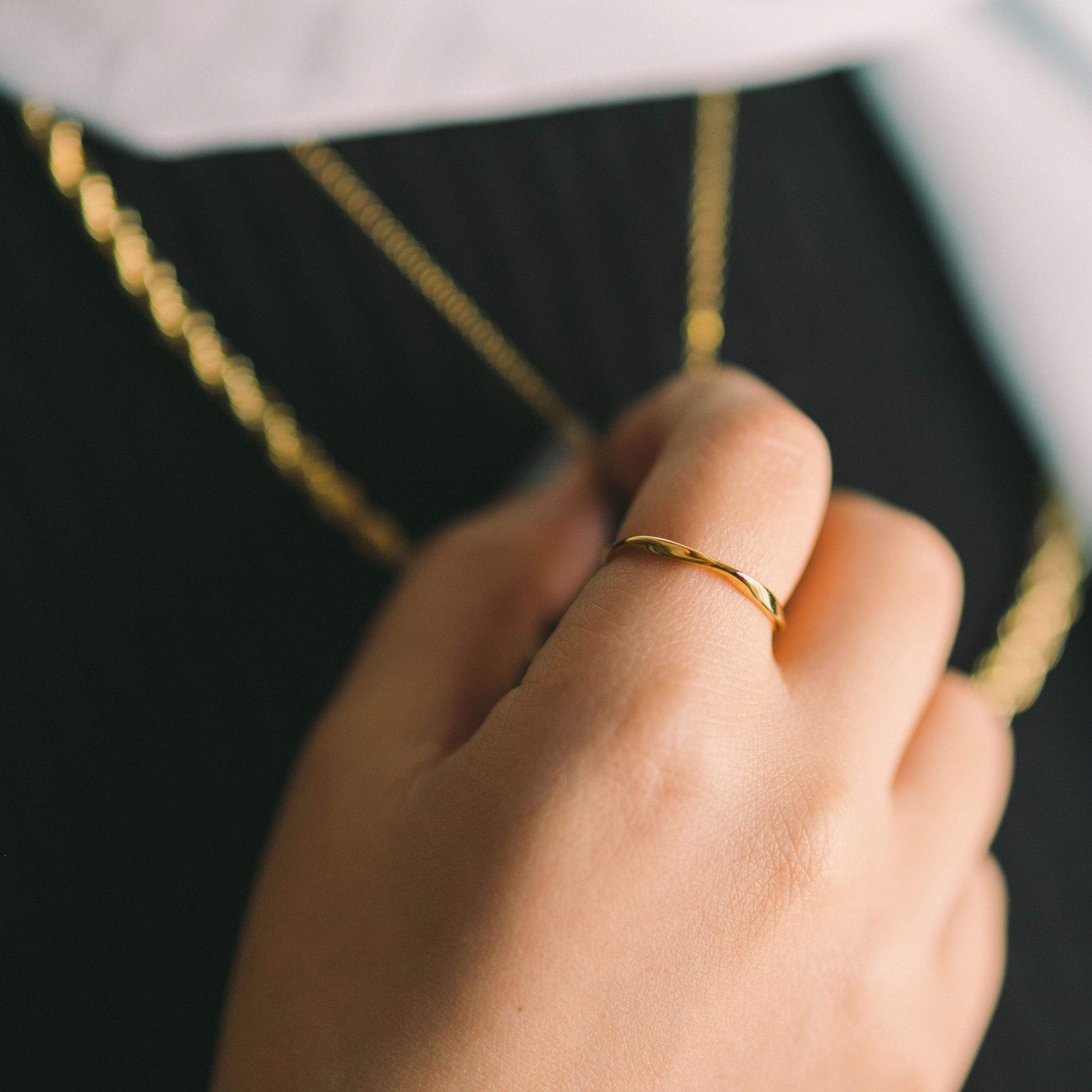 Close-up of a gold ripple ring worn on the index finger, with layered gold necklaces in the background.