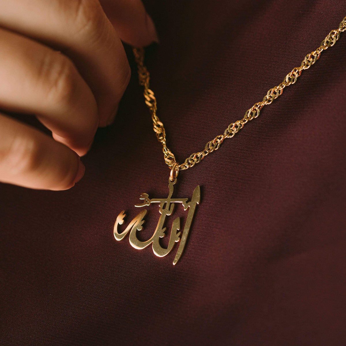 Close up of the gold Allah Pendant Necklace | Women against the brown fabric of the model's clothing.