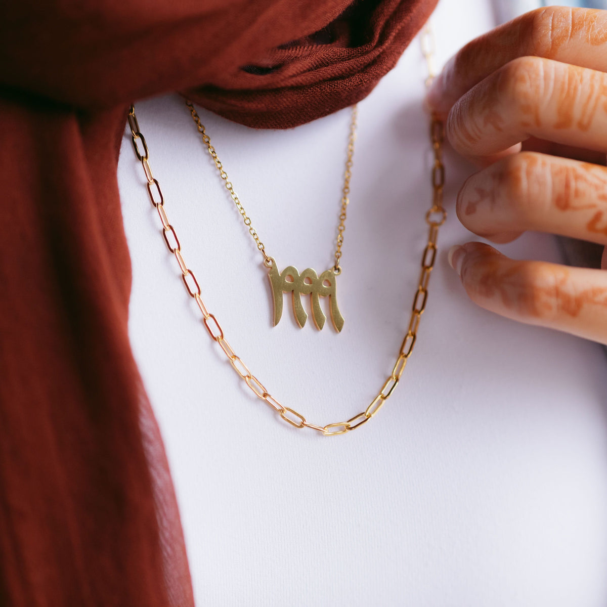 Gold link chain necklace layered with a gold Arabic numeral pendant necklace, styled with a maroon scarf and a hand with henna.
