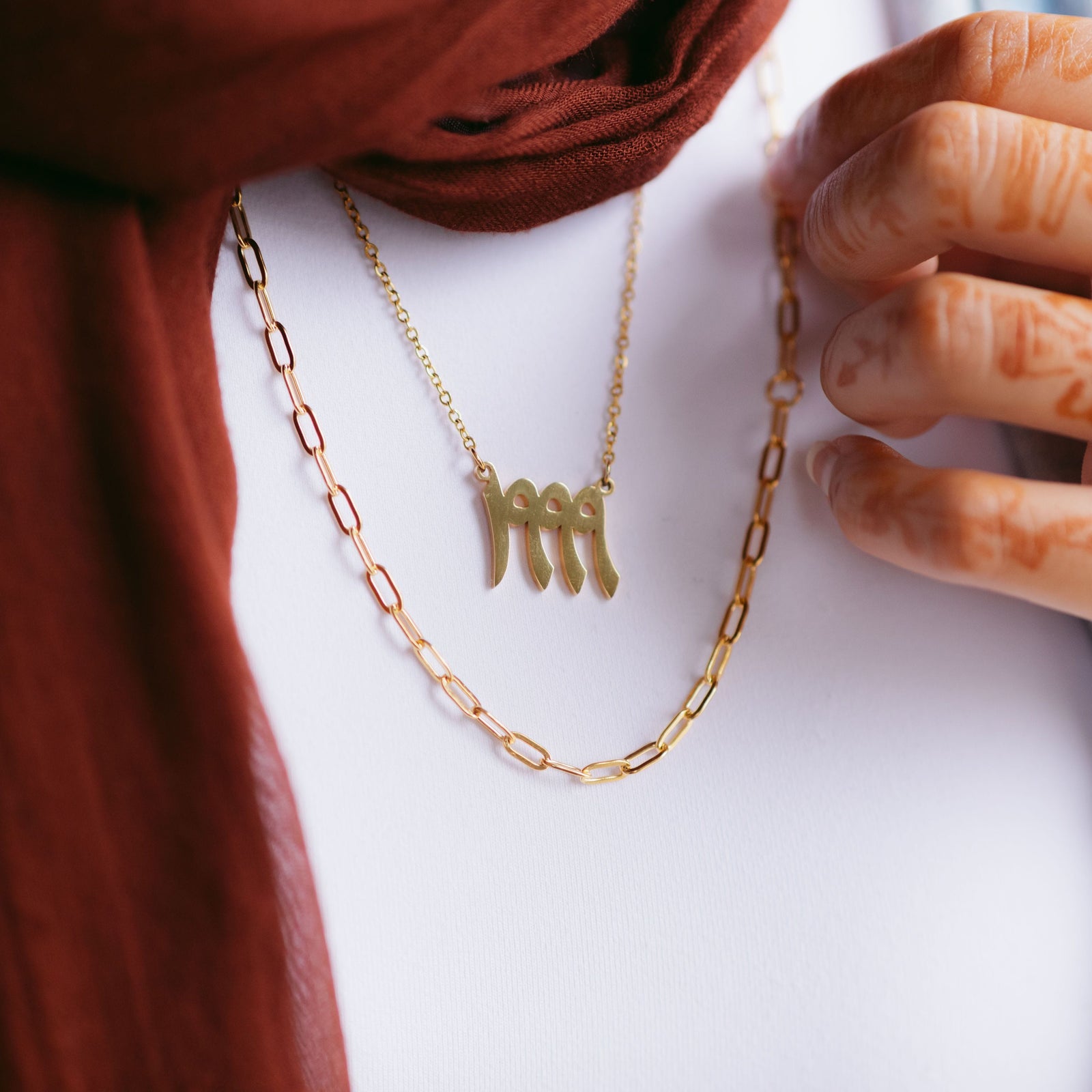 Gold link chain necklace layered with a gold Arabic numeral pendant necklace, styled with a maroon scarf and a hand with henna.