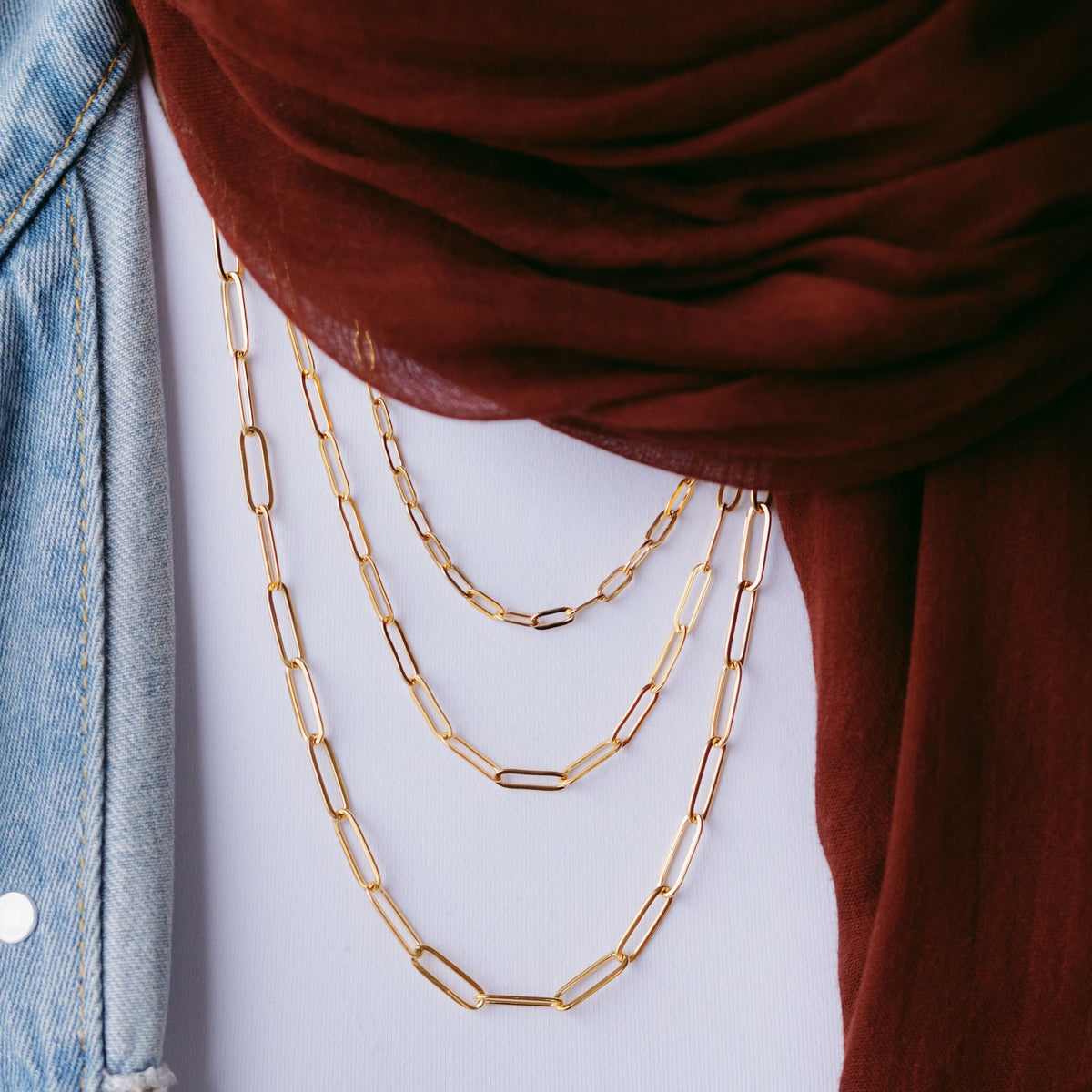Gold link chain necklaces layered over a white top, styled with a maroon scarf and light denim jacket.