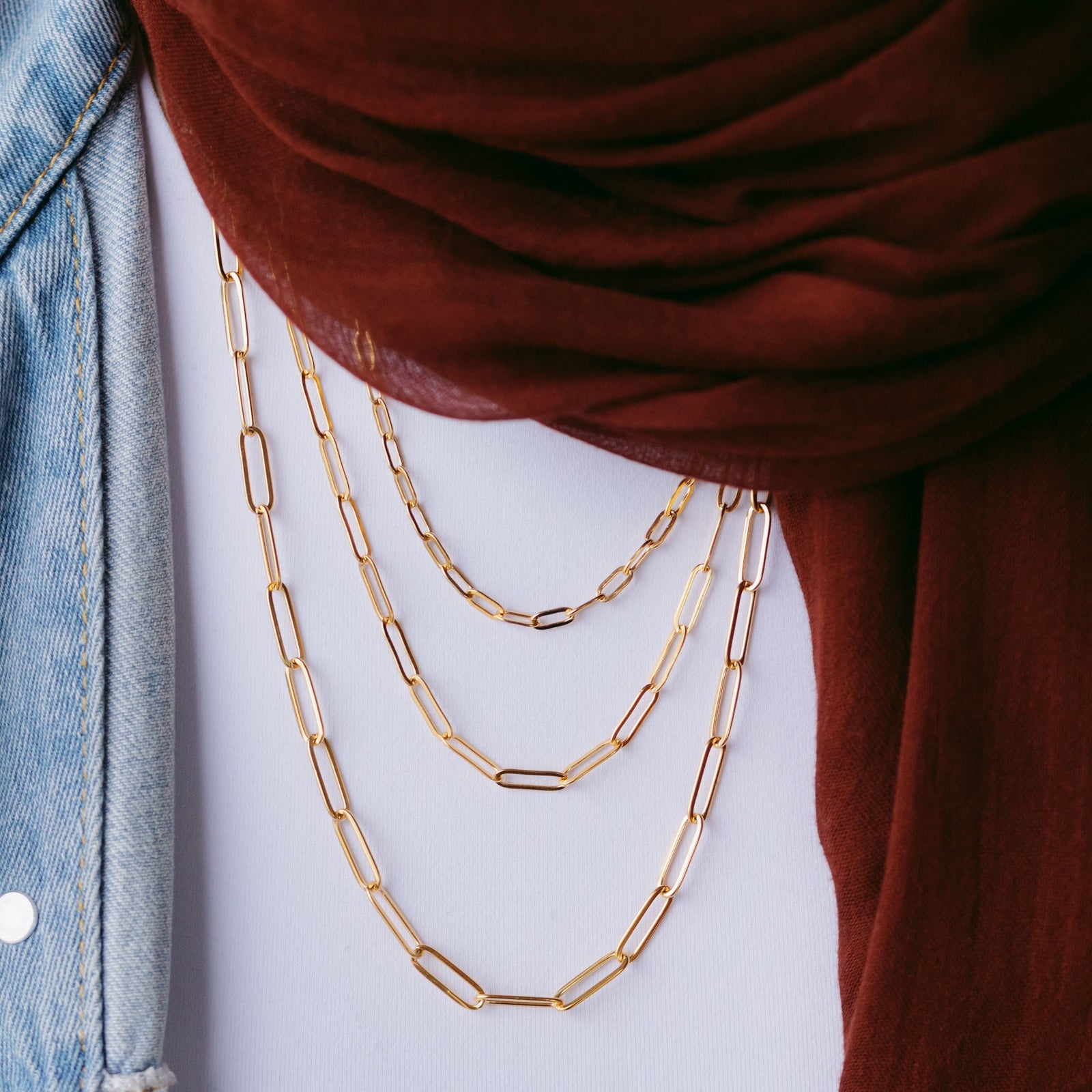 Gold link chain necklaces layered over a white top, styled with a maroon scarf and light denim jacket.