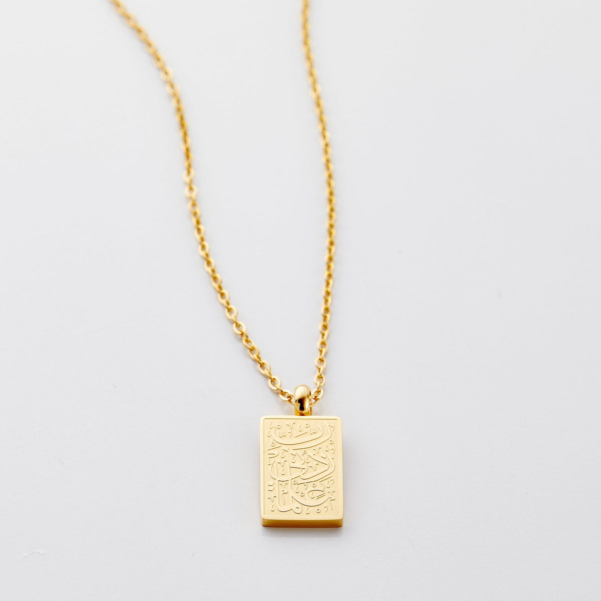 Flat lay of an 18k gold plated necklace with a small rectangular pendant that features Arabic calligraphy that translates to "My Lord, increase me in knowledge."