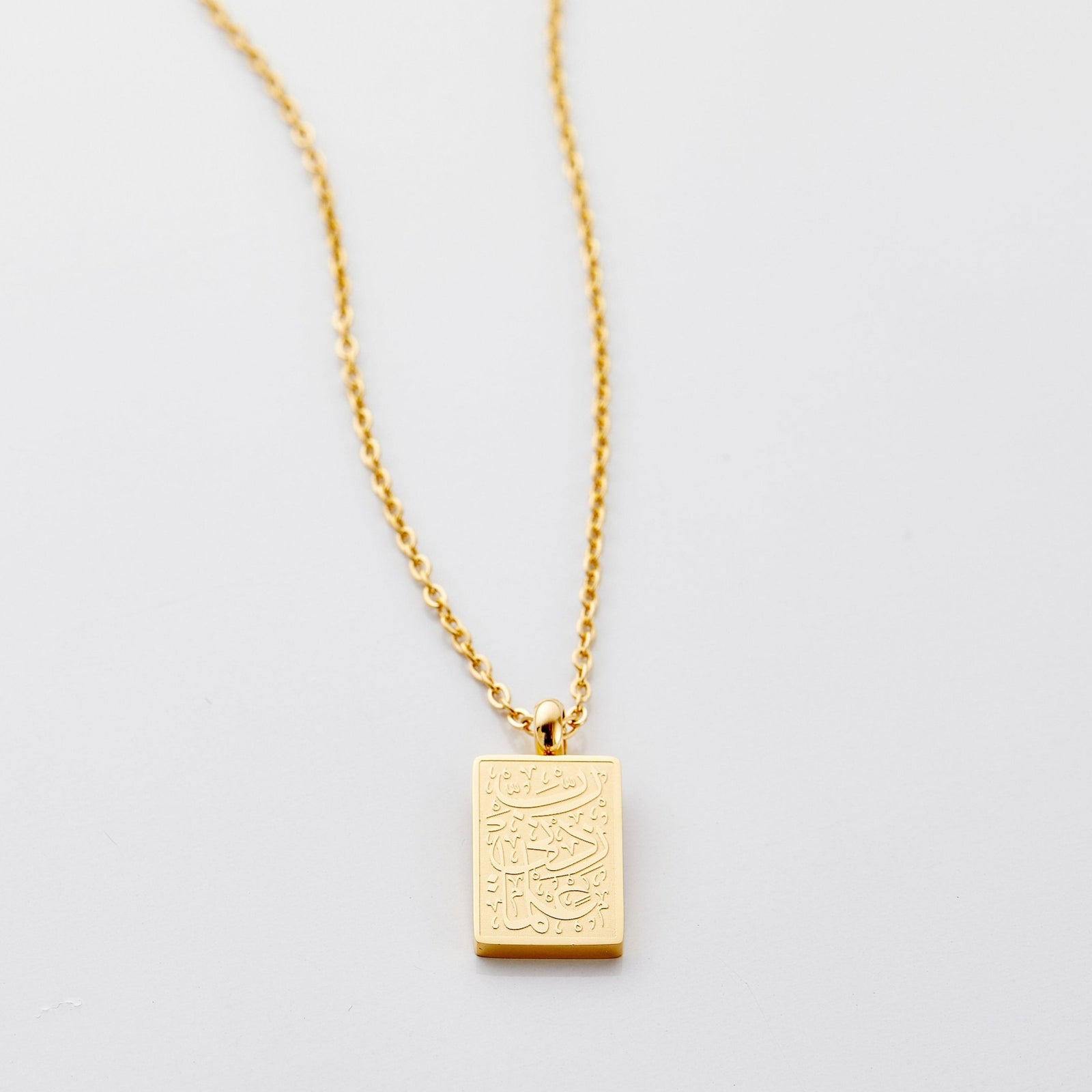 Flat lay of an 18k gold plated necklace with a small rectangular pendant that features Arabic calligraphy that translates to "My Lord, increase me in knowledge."