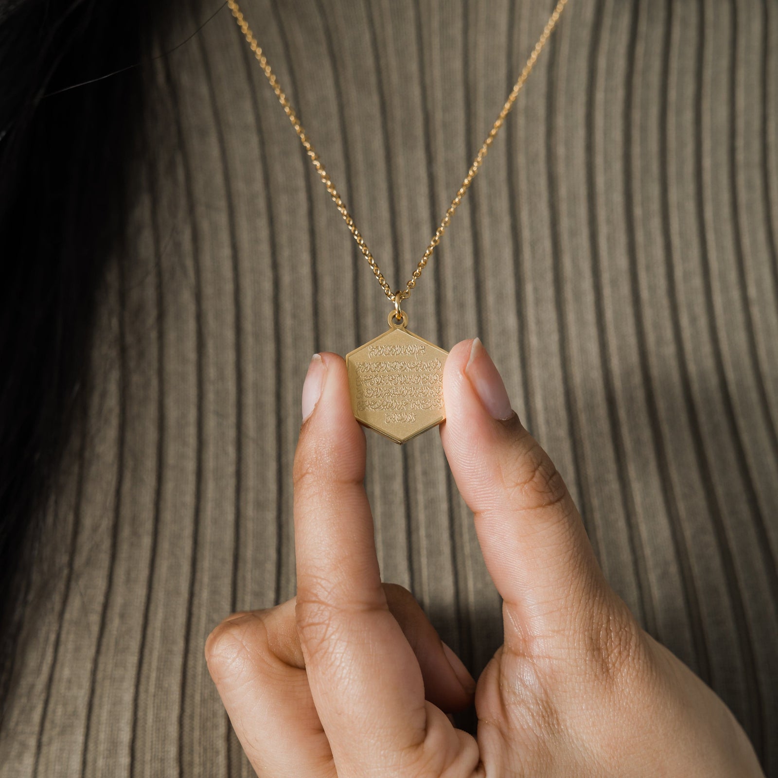 Close up of the gold Surat Al-Fatiha Necklace as the model holds its pendant between their fingers.