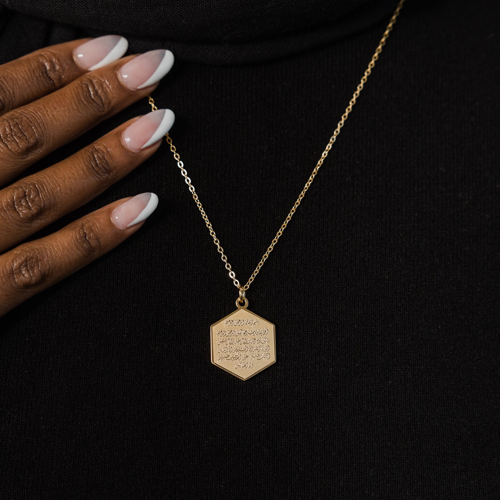 Close up of the gold Surat Al-Fatiha Necklace against the black fabric of the model's clothing.