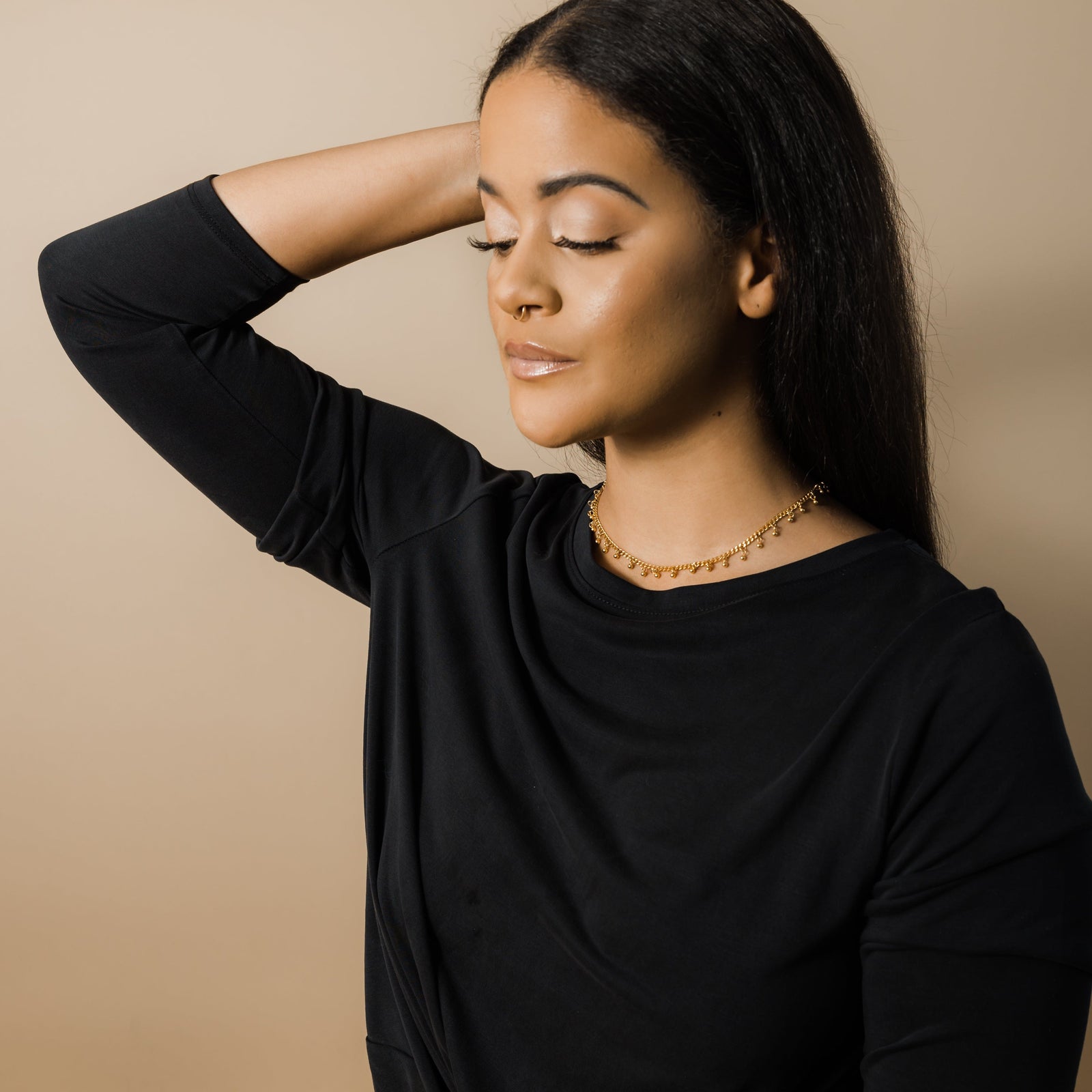 Woman wearing a gold choker with dangling beads, dressed in a black top, arm raised behind head.