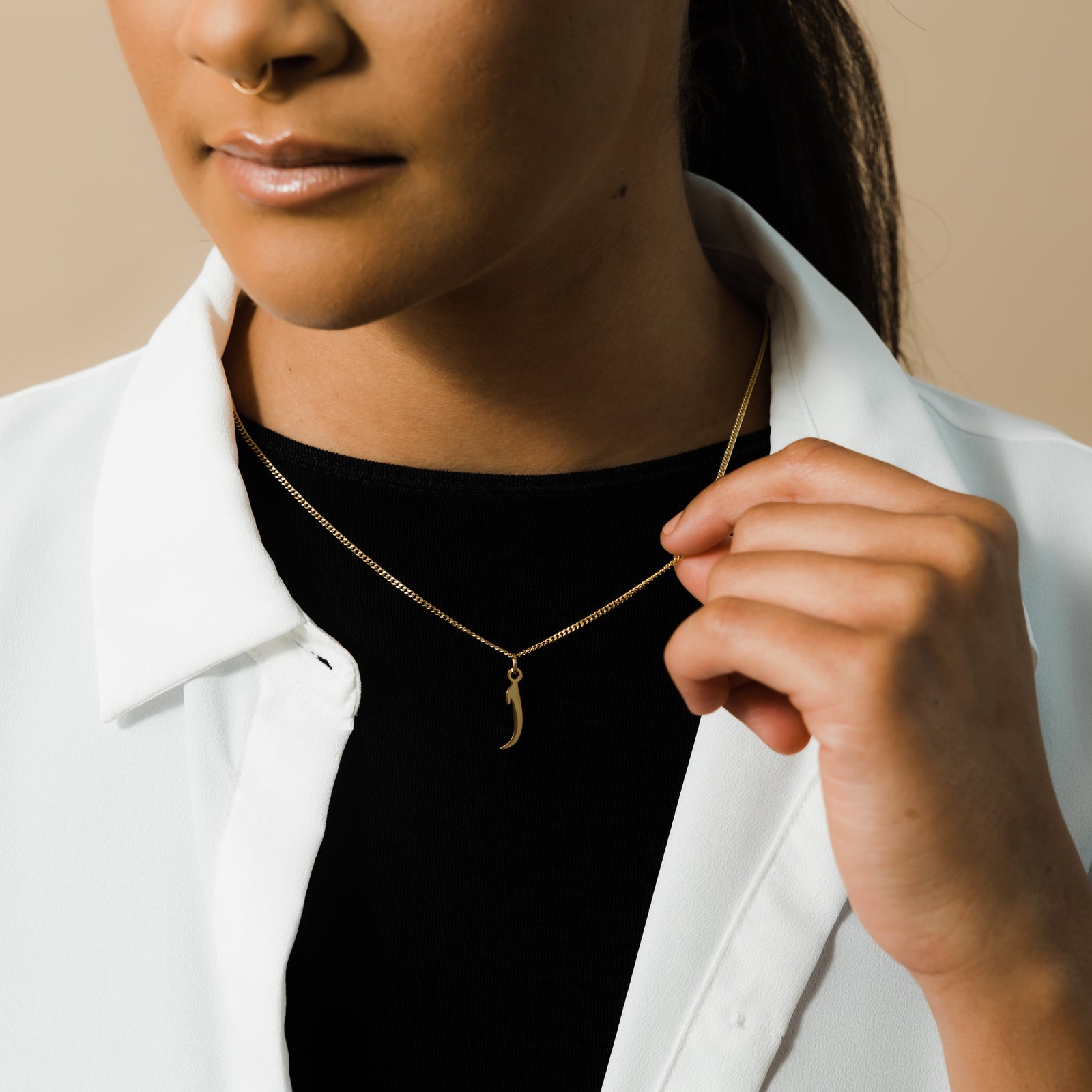Close up of the gold Hanging Arabic Letter Necklace as worn by the model. The model lifts the chain to show is design.