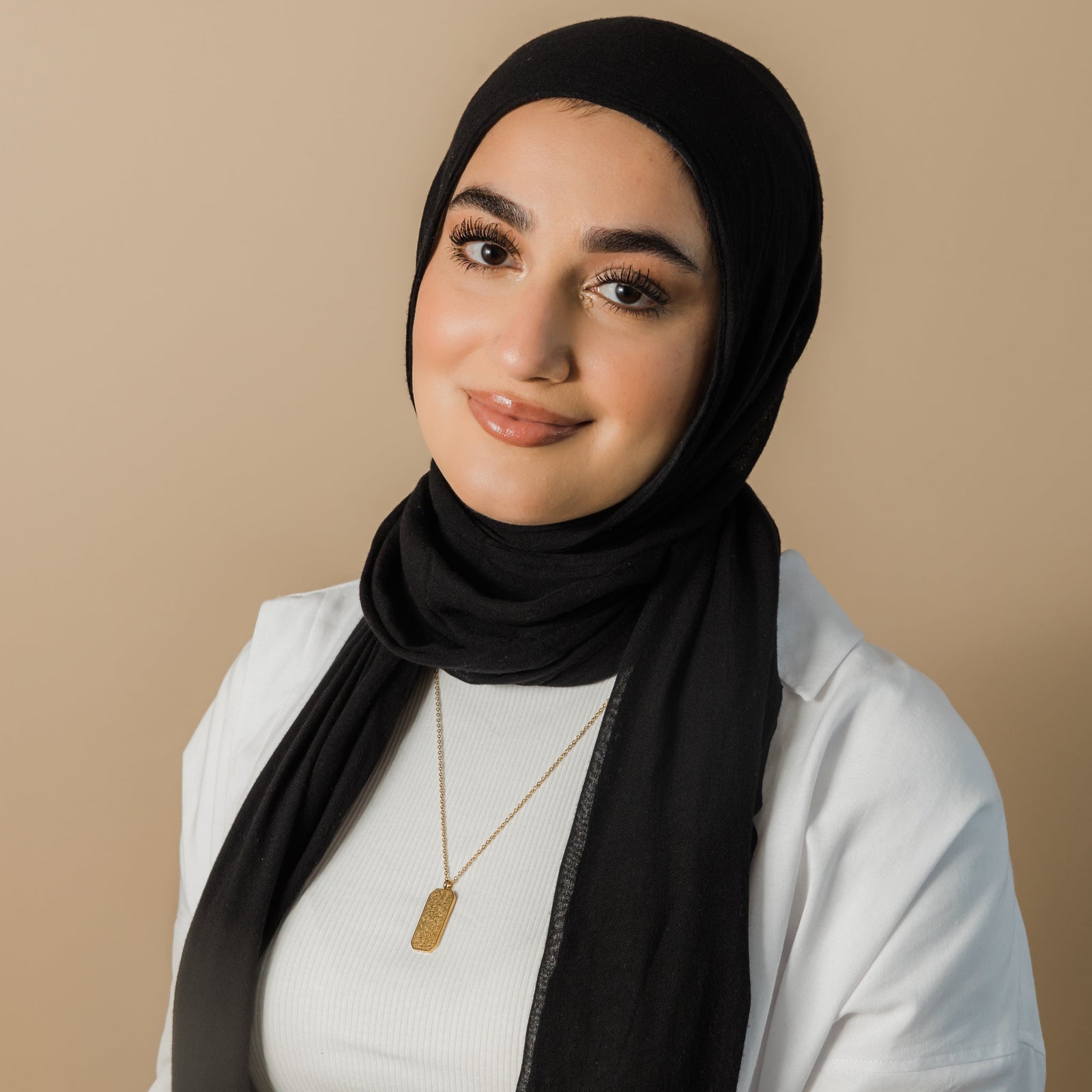 Photo of the model smiling to the camera. The model is wearing a black colored hijab, white shirt, and the gold Ayatul Kursi Tag necklace around their neck.