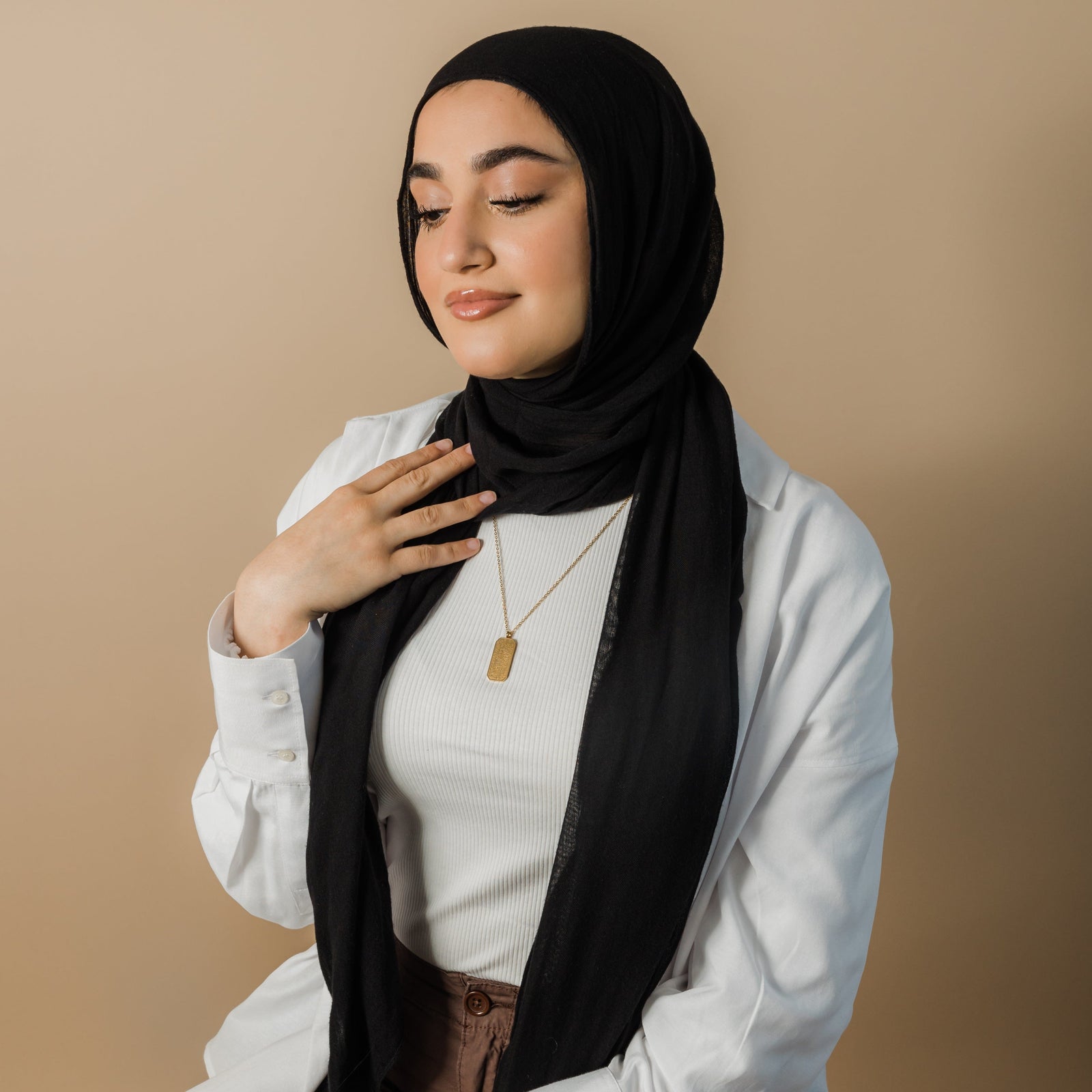 Photo of the model looking down, away from the camera, touching the fabric of their hijab. The model is wearing the gold Aytul Kursi.